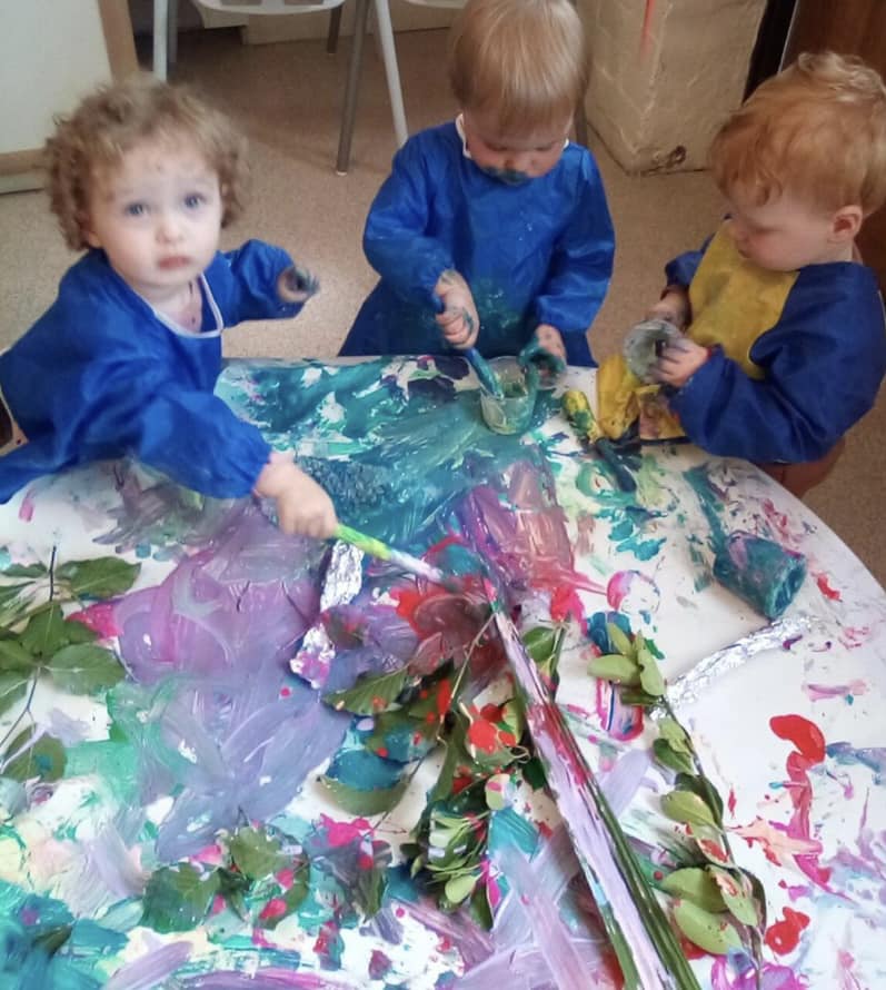 Children painting with various colors on a table; leaves and brushes used; blue smocks worn; messy and colorful.