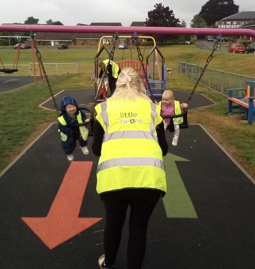 Adult pushing two children on swings at a playground; all wear yellow vests.