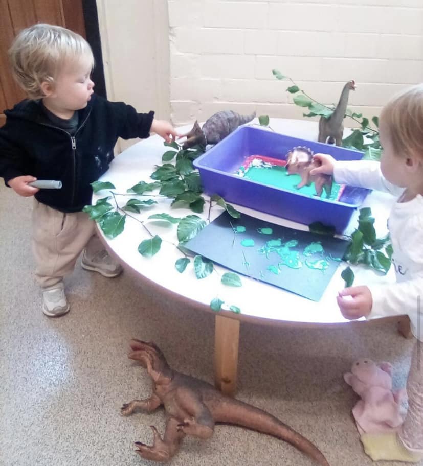 Two toddlers playing with dinosaur toys on a table with greenery. One points, the other reaches for a dino.