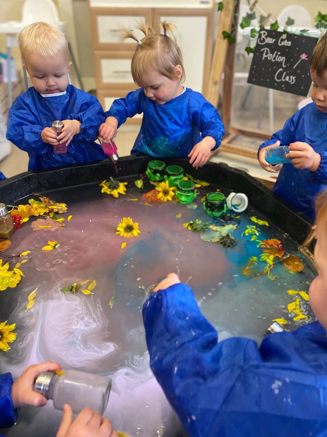Children in blue smocks playing with colorful liquids and flowers in a sensory table.