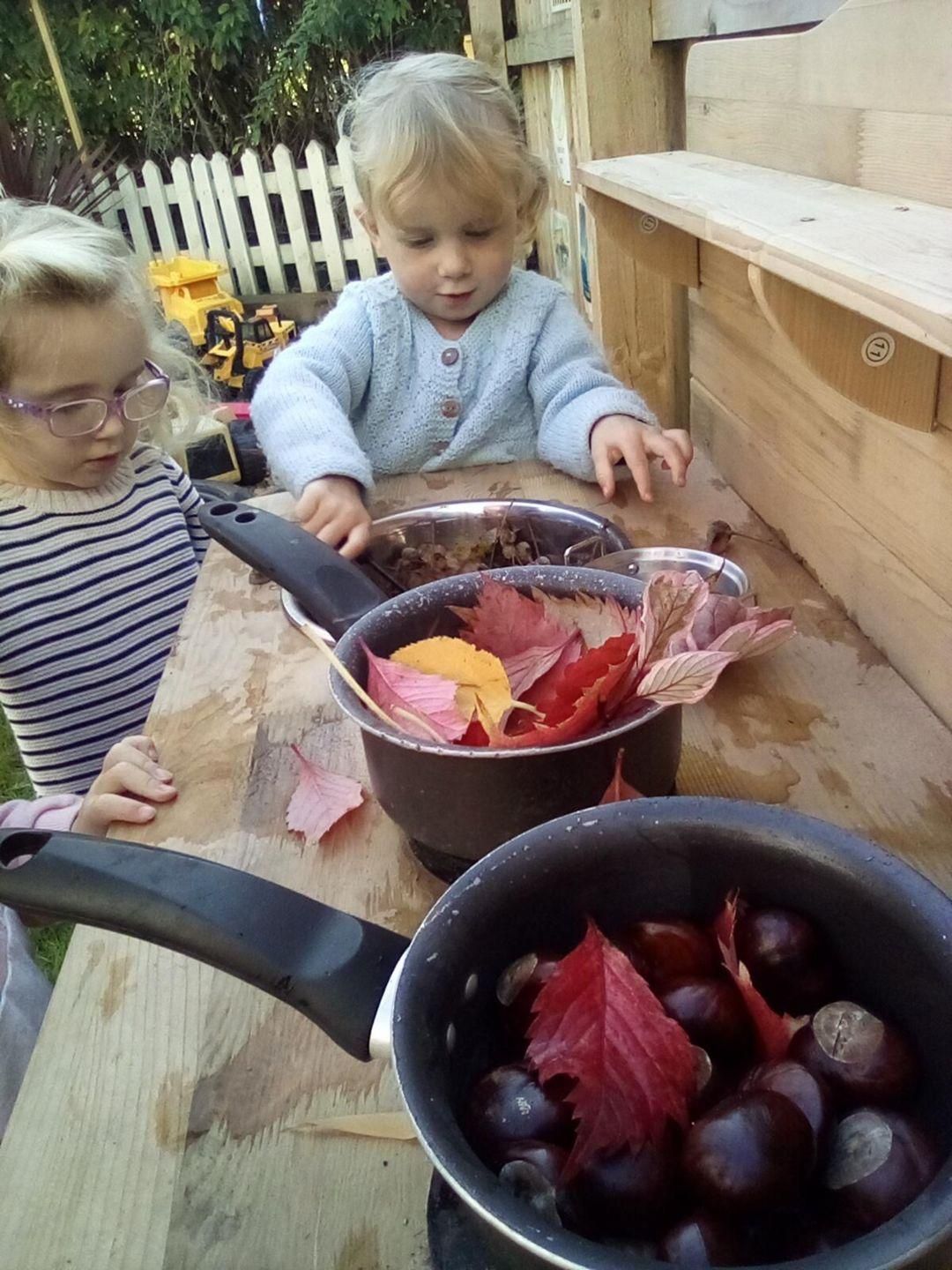 Two children playing with leaves and chestnuts in pots outdoors.