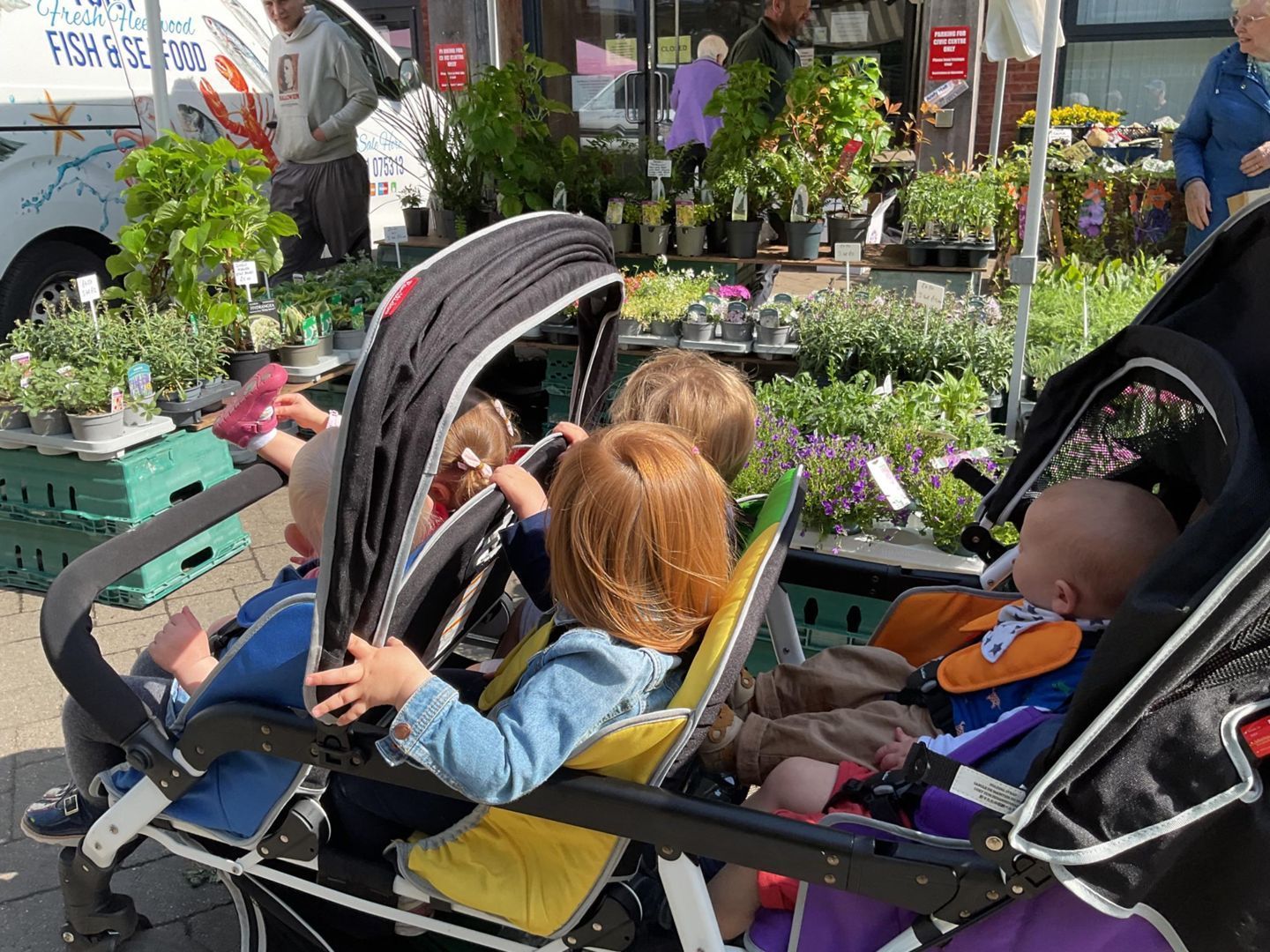Four children in a double stroller at a plant market. Plants and people in the background.