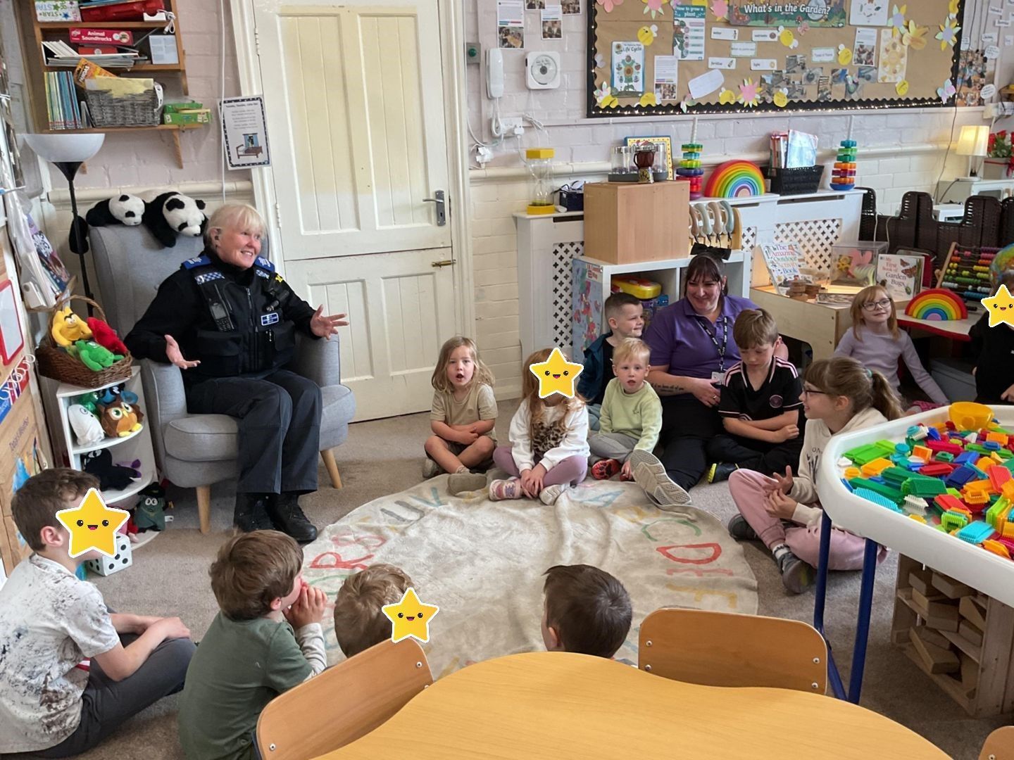A person sits reading to children in a classroom. Kids sit on a floor mat, smiling. Colorful toys are visible.