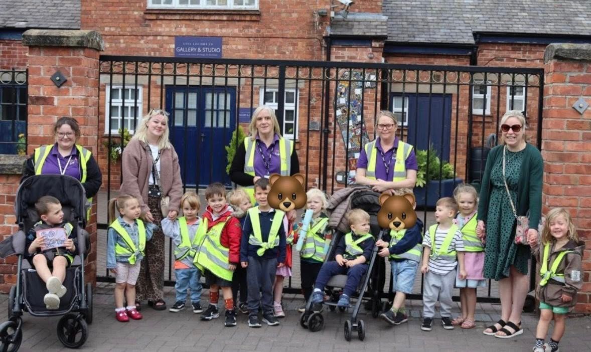 Group of young children and adults outside building with iron gate; children wearing vests.