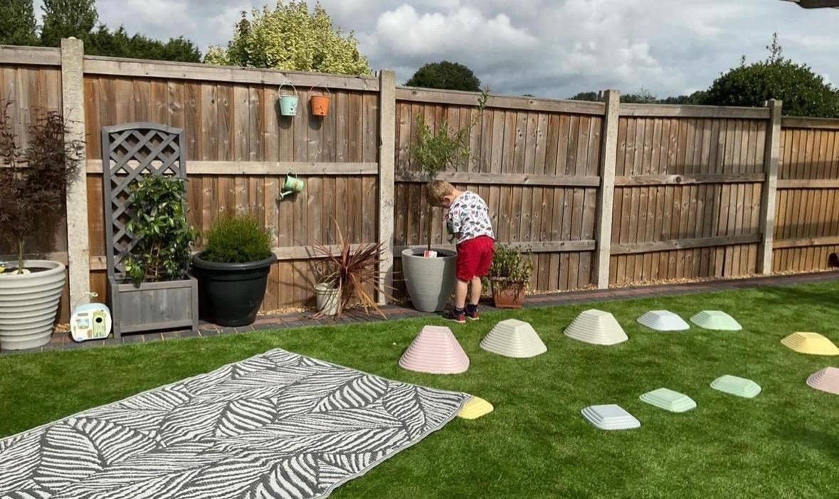 Child exploring a plant in a backyard. A zebra-striped mat and colored cones are on the grass. Wooden fence in background.