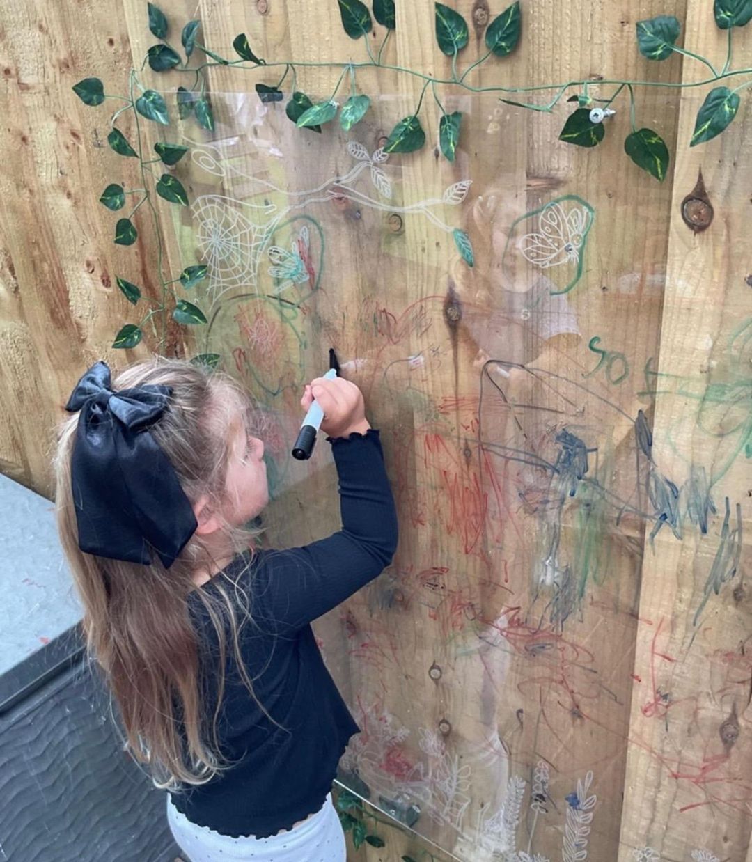 Girl with a black bow draws on a wooden fence decorated with fake leaves.