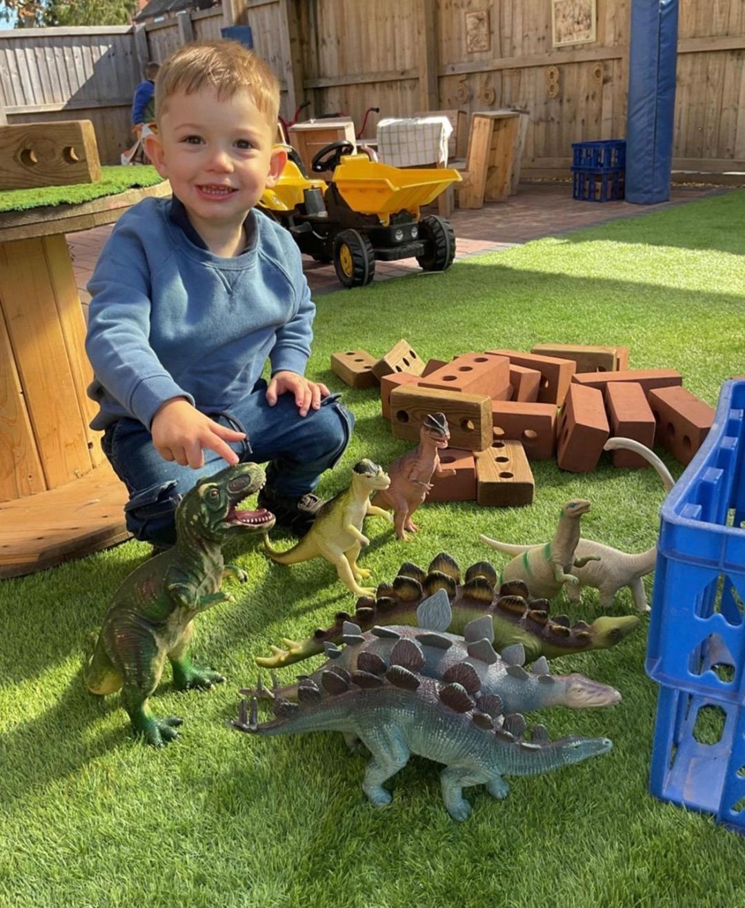 Boy smiles, pointing at toy dinosaurs on green grass. Bricks and blue crate are nearby.