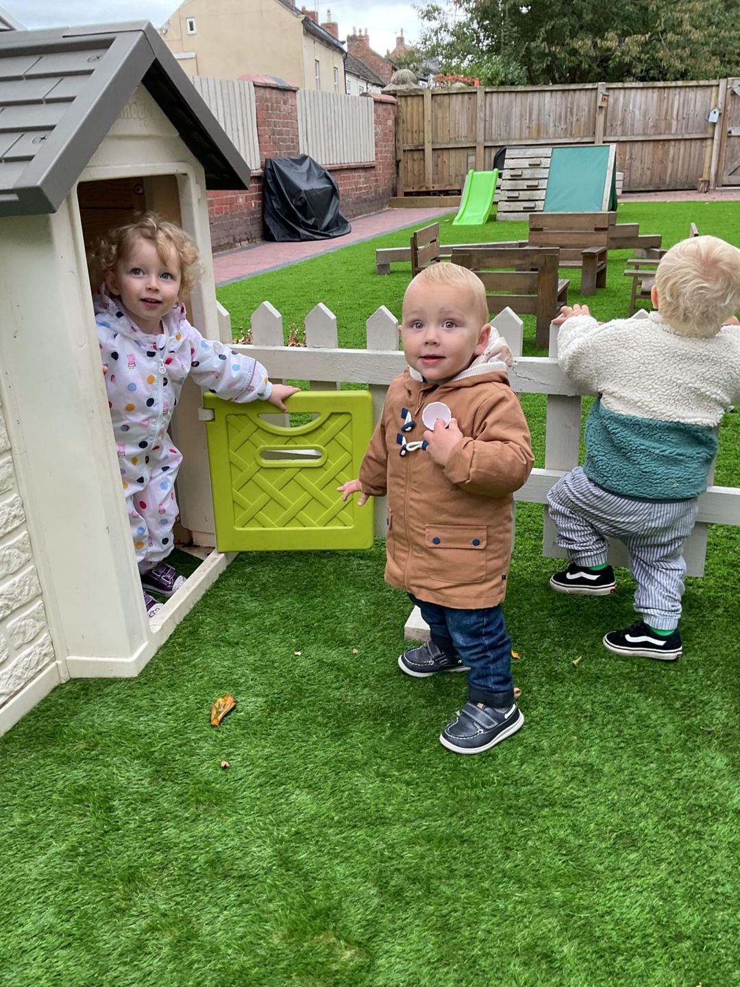 Three toddlers playing outdoors: one in a playhouse, one in a jacket, and one climbing a fence.