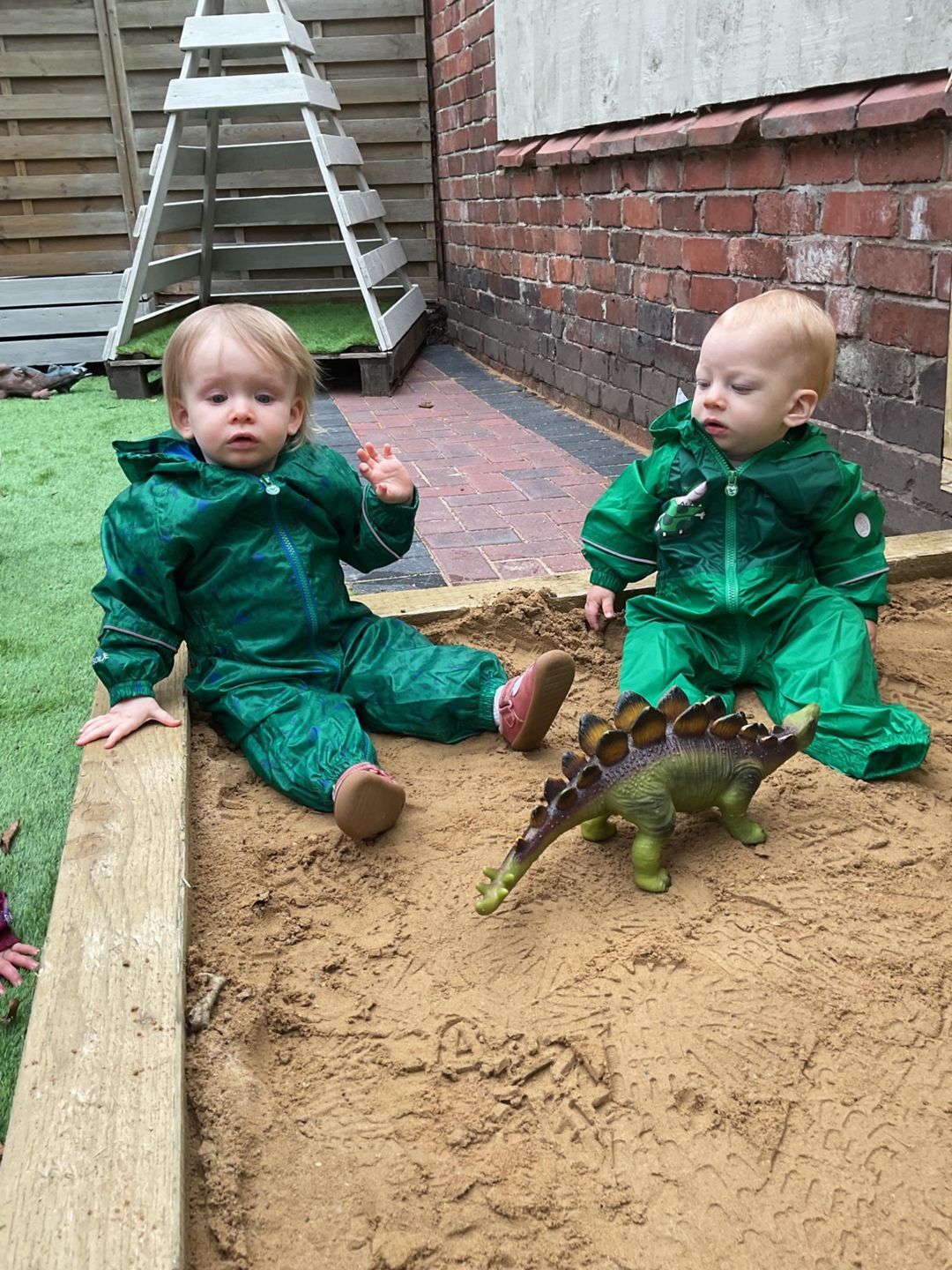 Two toddlers in green rainsuits play in a sandbox with a dinosaur toy.
