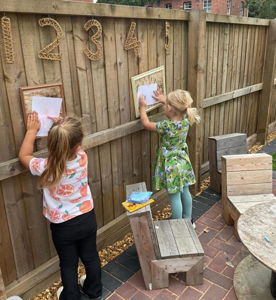 Two children at wooden fence, placing paper in frames beneath numbers. Outdoor setting with small furniture.