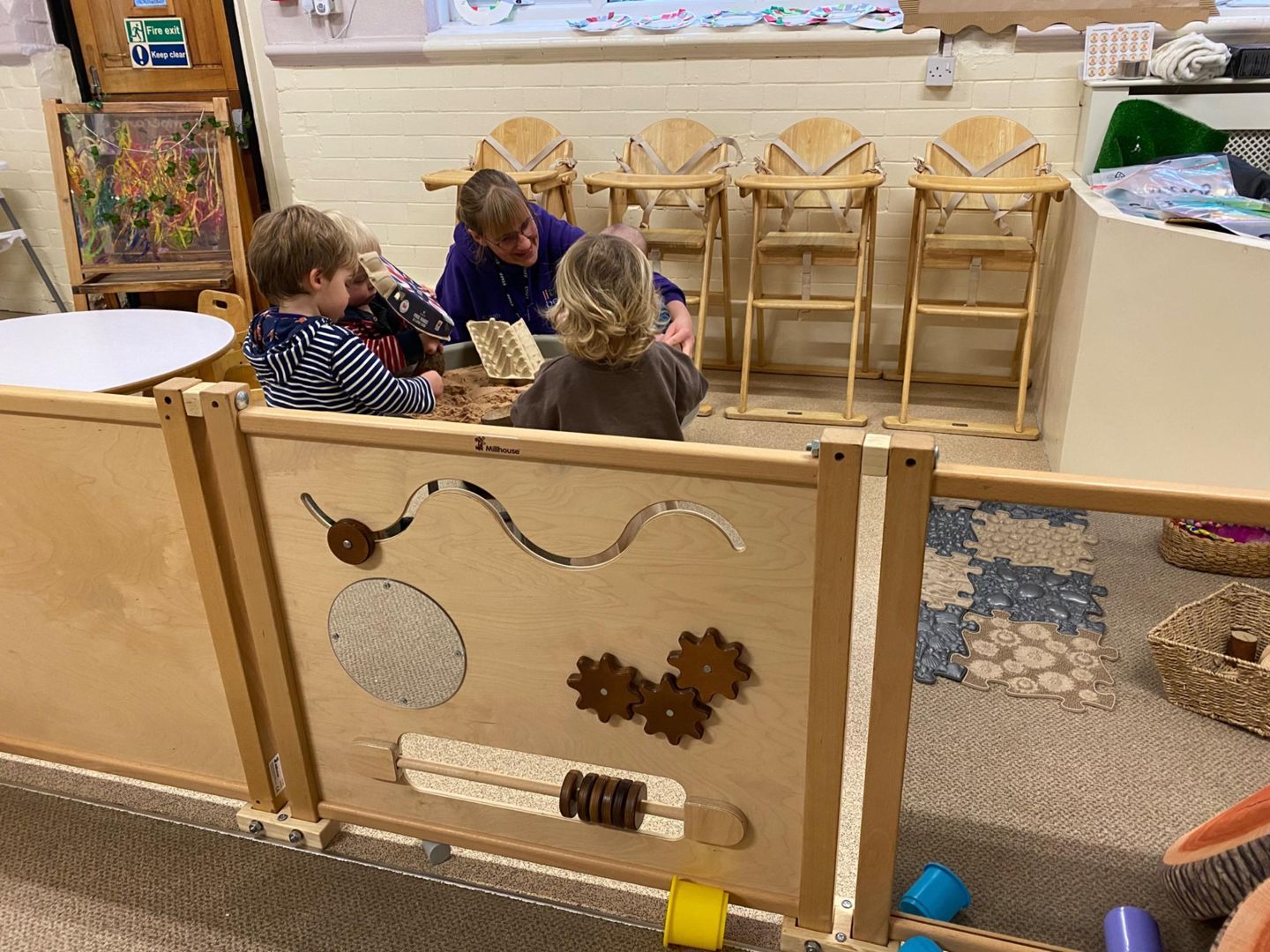Three children and an adult looking at a book in a nursery room. Sensory panel in the foreground.