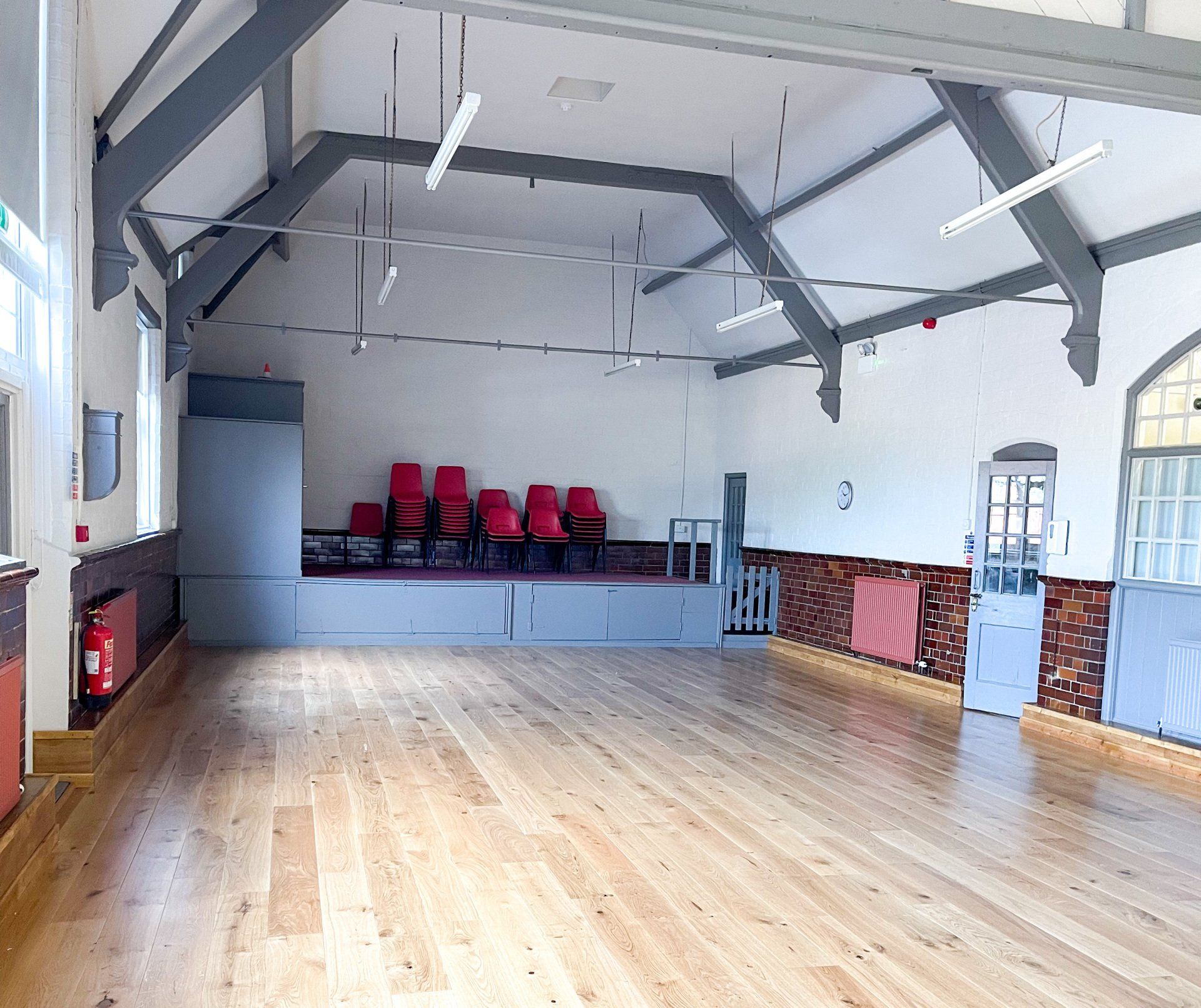 Empty hall with wood floor, stage with red chairs, and high ceiling with beams.