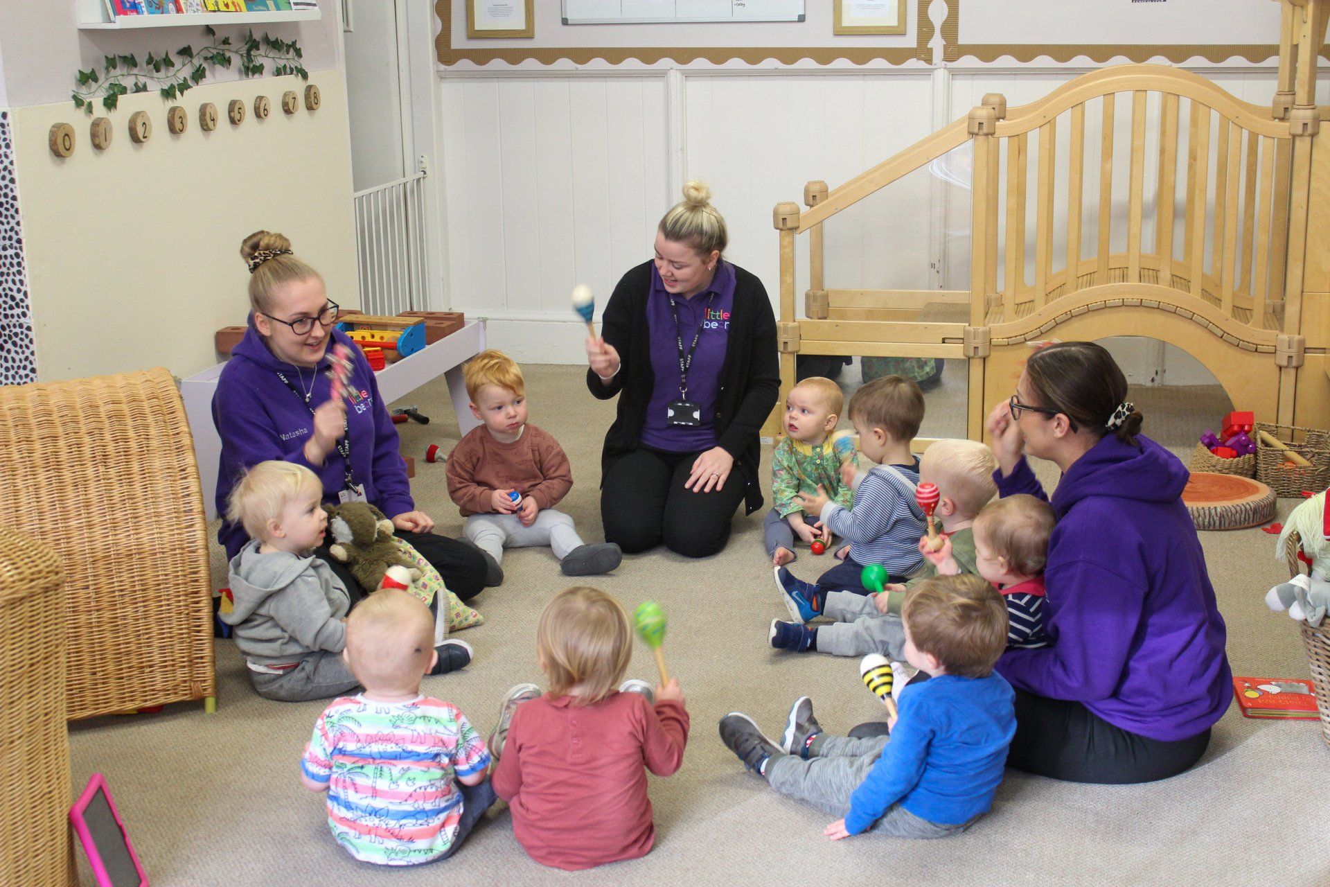 Nursery staff lead children in a circle, playing musical instruments. They are smiling in a playroom setting.