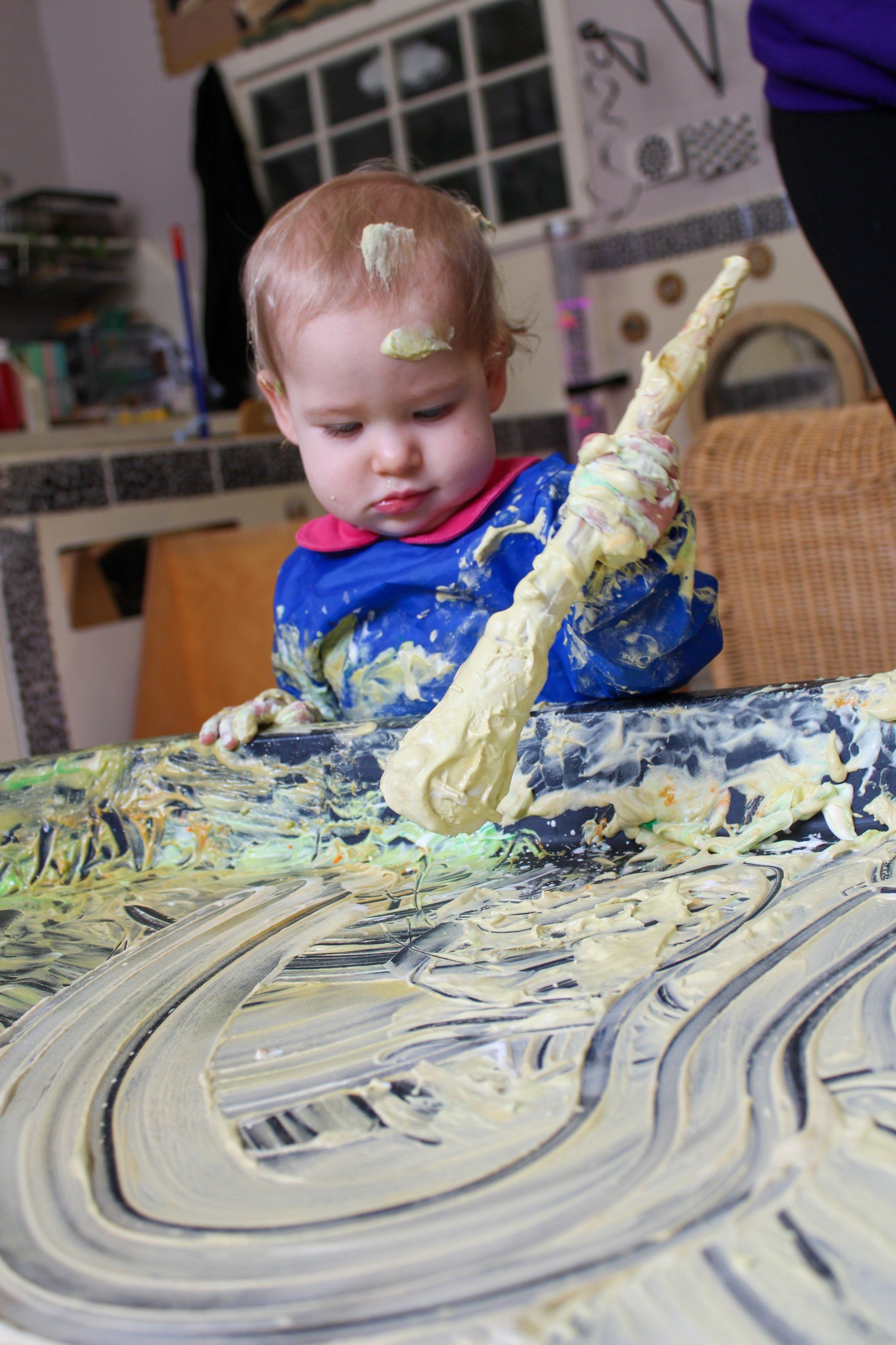 Child covered in frosting, smearing it on a table with a spatula.