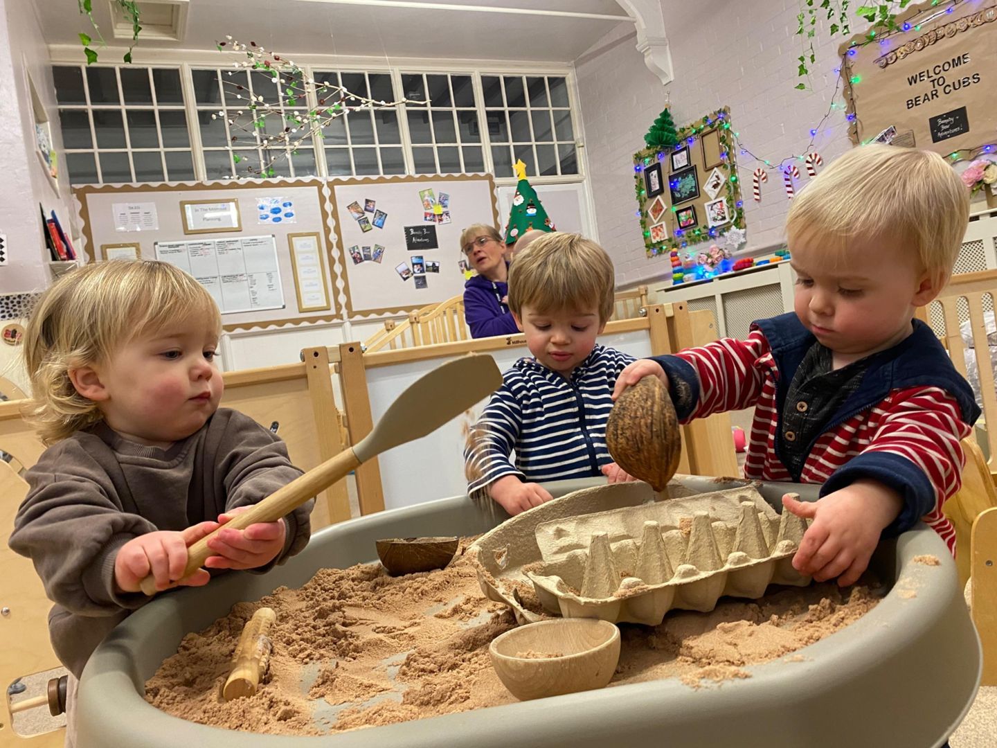 Three toddlers playing with sand in a sensory table. One holds a wooden spoon, another a coconut. Bright, indoor setting.