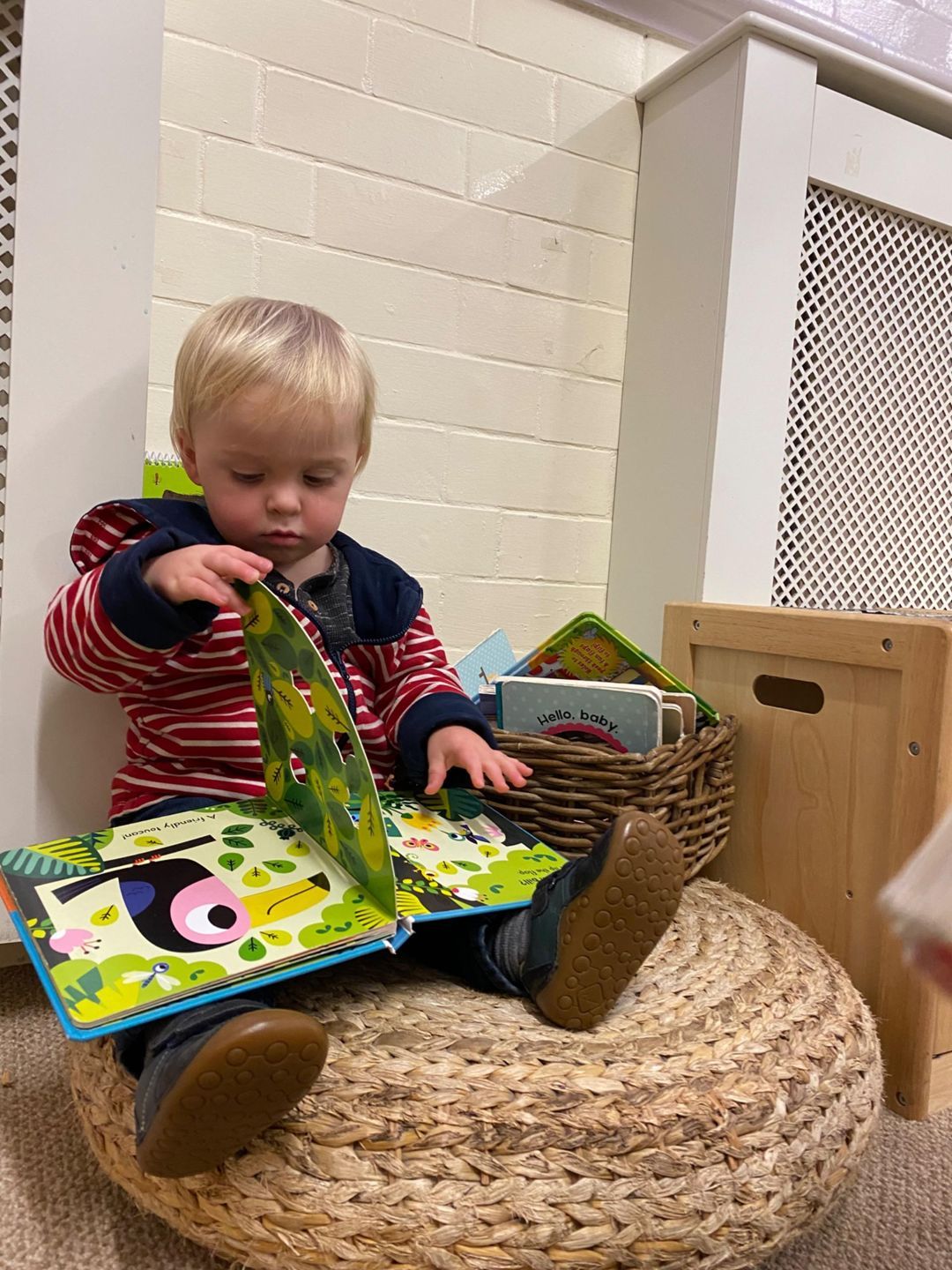 Child seated reading a book, other books nearby in a basket. Red striped top, sitting on a woven pouf.