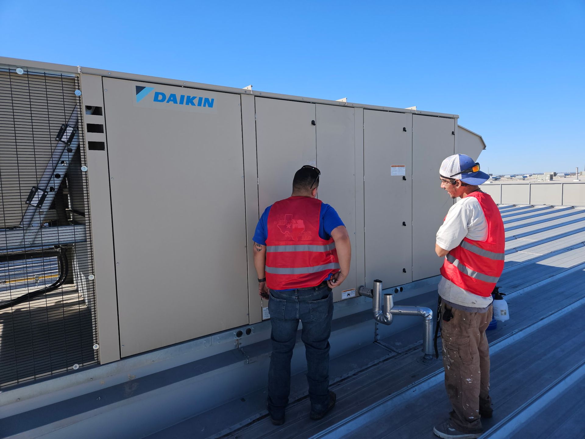 Two workers in red vests inspect a Daikin rooftop HVAC unit on a sunny day.