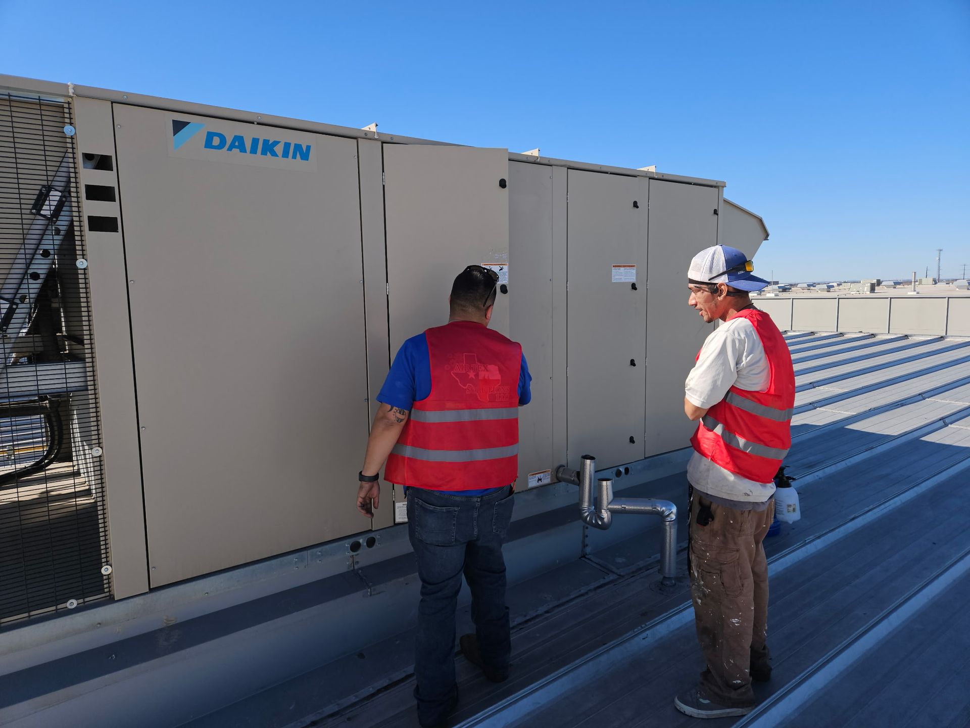 Two workers in red vests on a roof inspecting a Daikin HVAC unit on a sunny day.