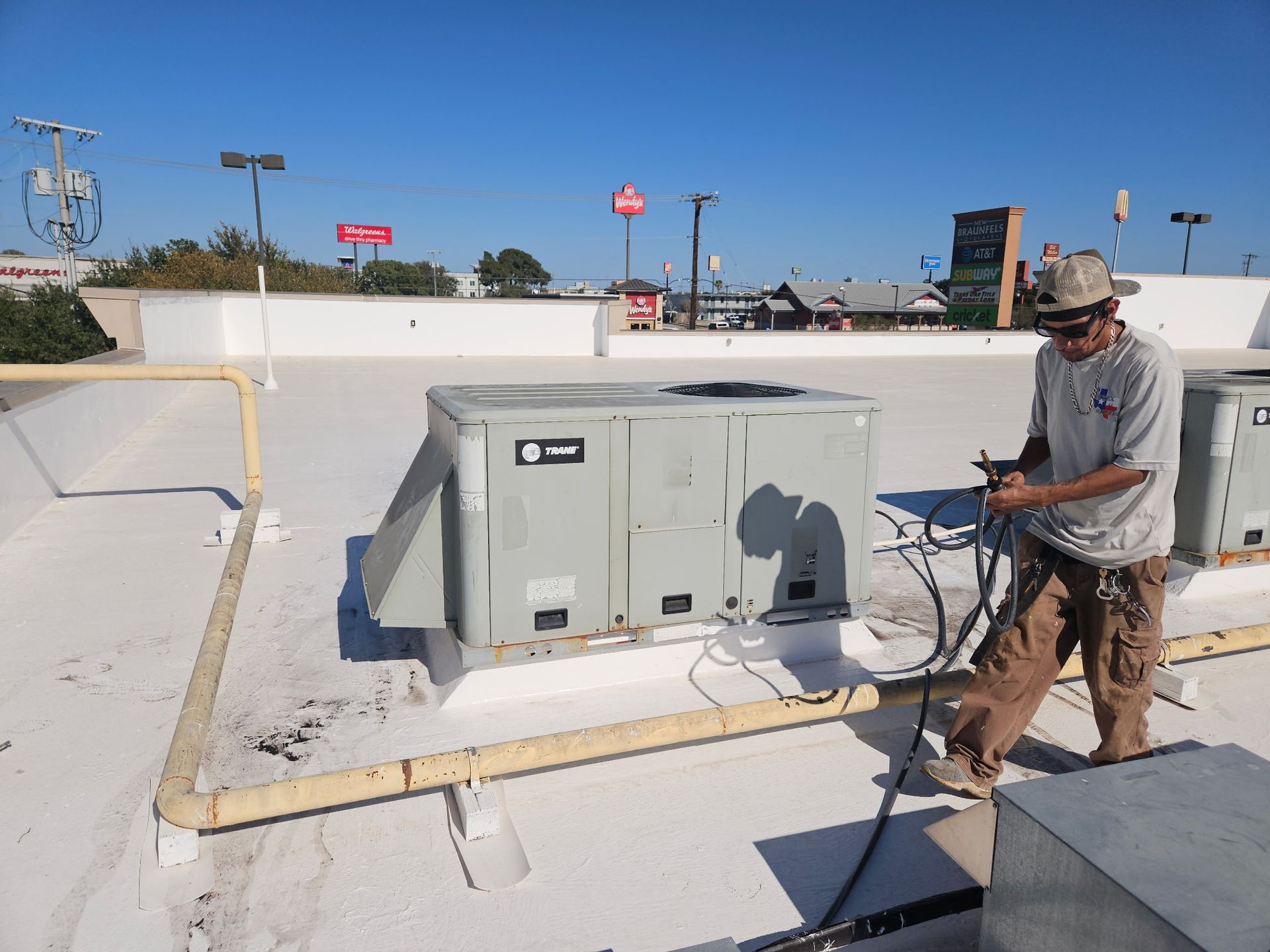 Man on roof works on HVAC unit; yellow pipes, white roof.