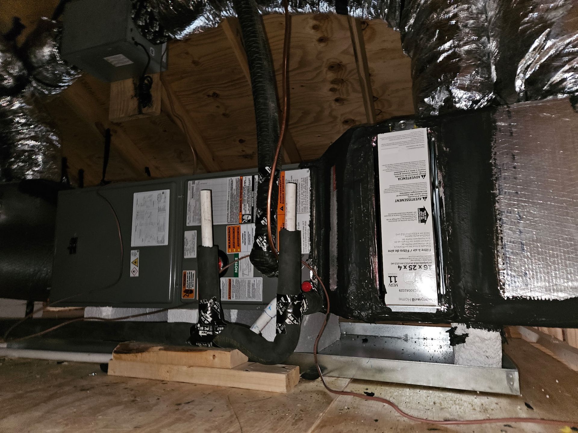 HVAC unit in attic, black ductwork, silver insulation, mounted on wooden supports.