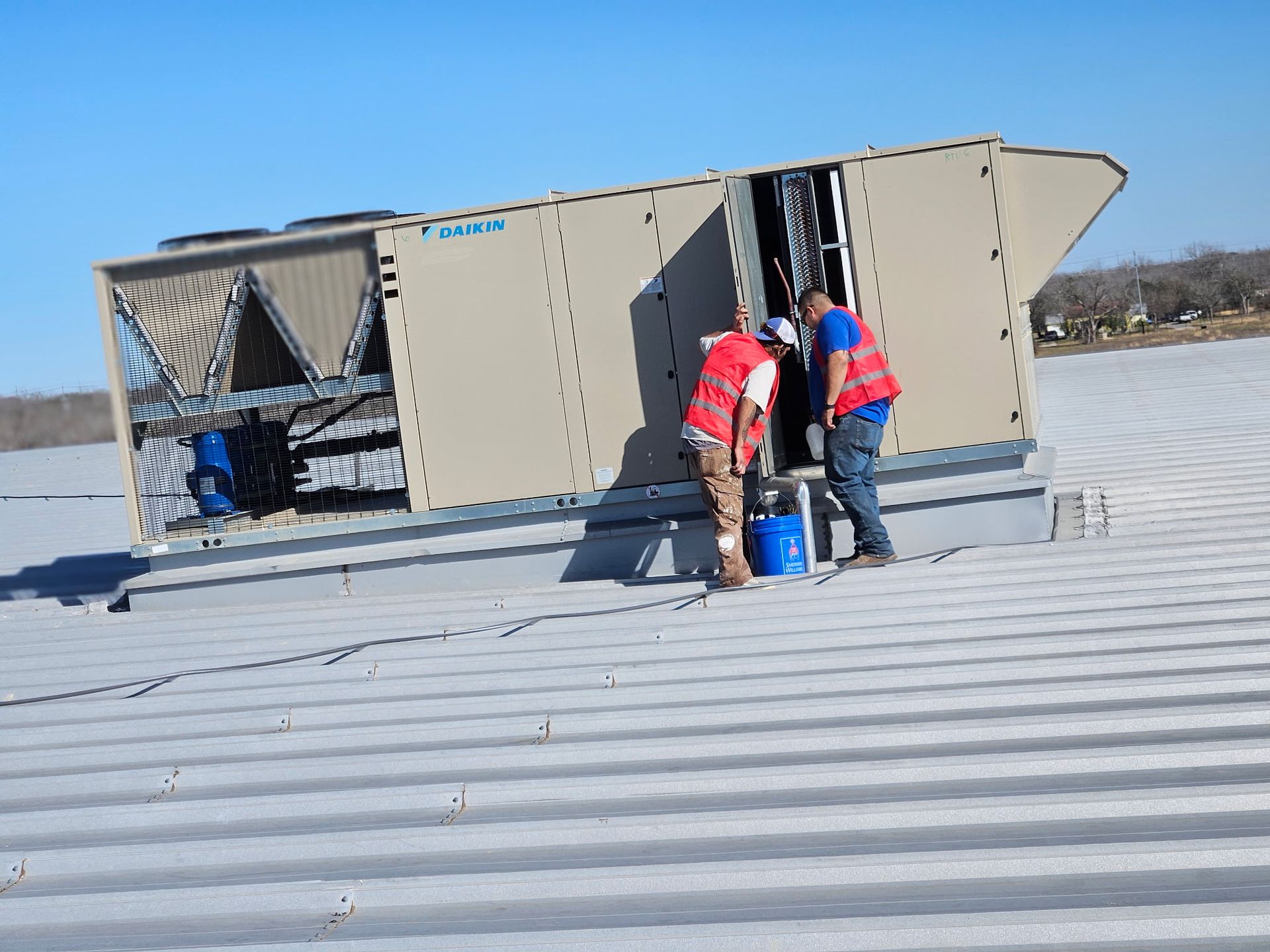 Two people in safety vests inspect an HVAC unit on a rooftop under a clear sky.