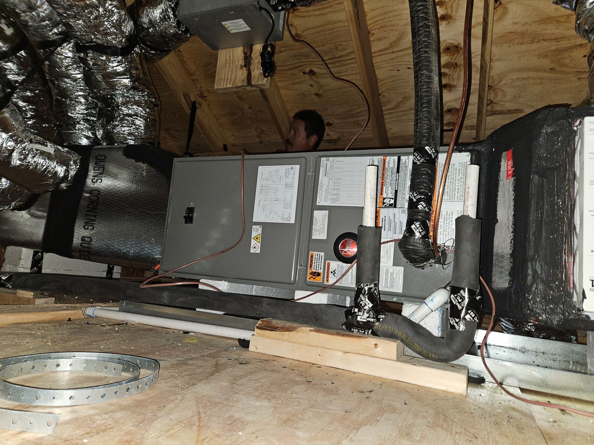 Person working on HVAC unit in attic, gray metal box, ductwork, wood supports, exposed insulation.