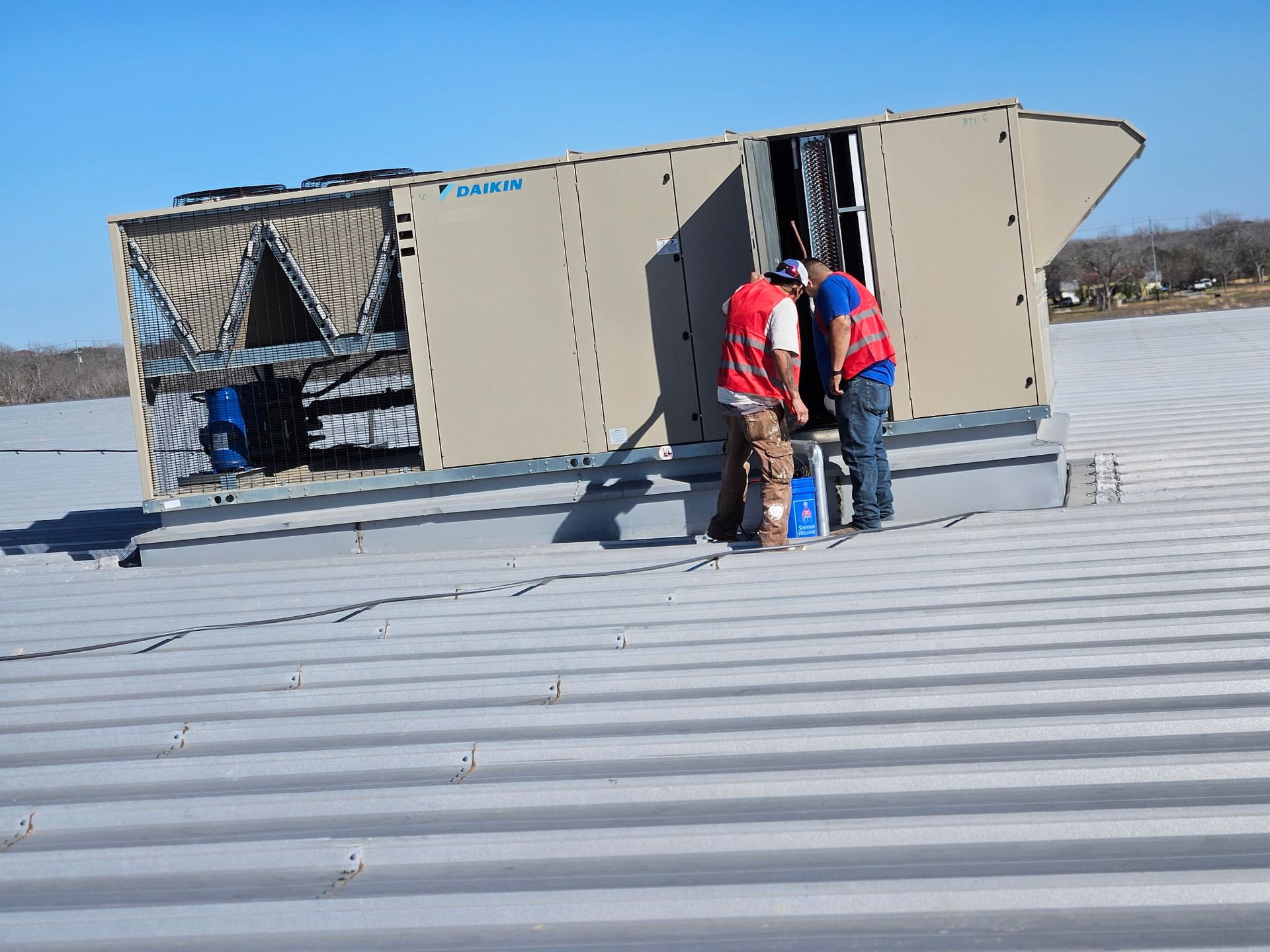 Two workers inspecting rooftop HVAC unit. Silver roof, blue sky, tan equipment.