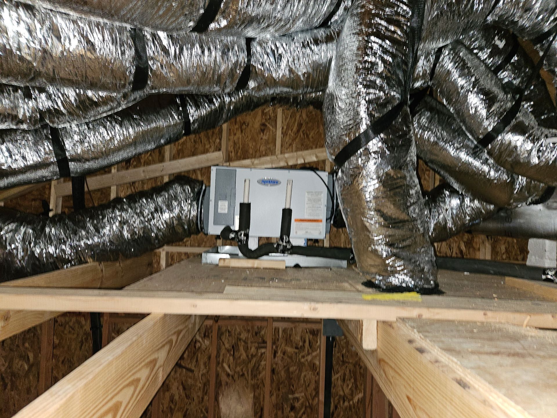 HVAC unit in an attic with insulated ductwork. Wooden beams and plywood flooring support the equipment.