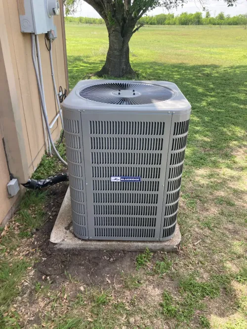 A gray air conditioner is sitting on top of a concrete platform next to a tree.