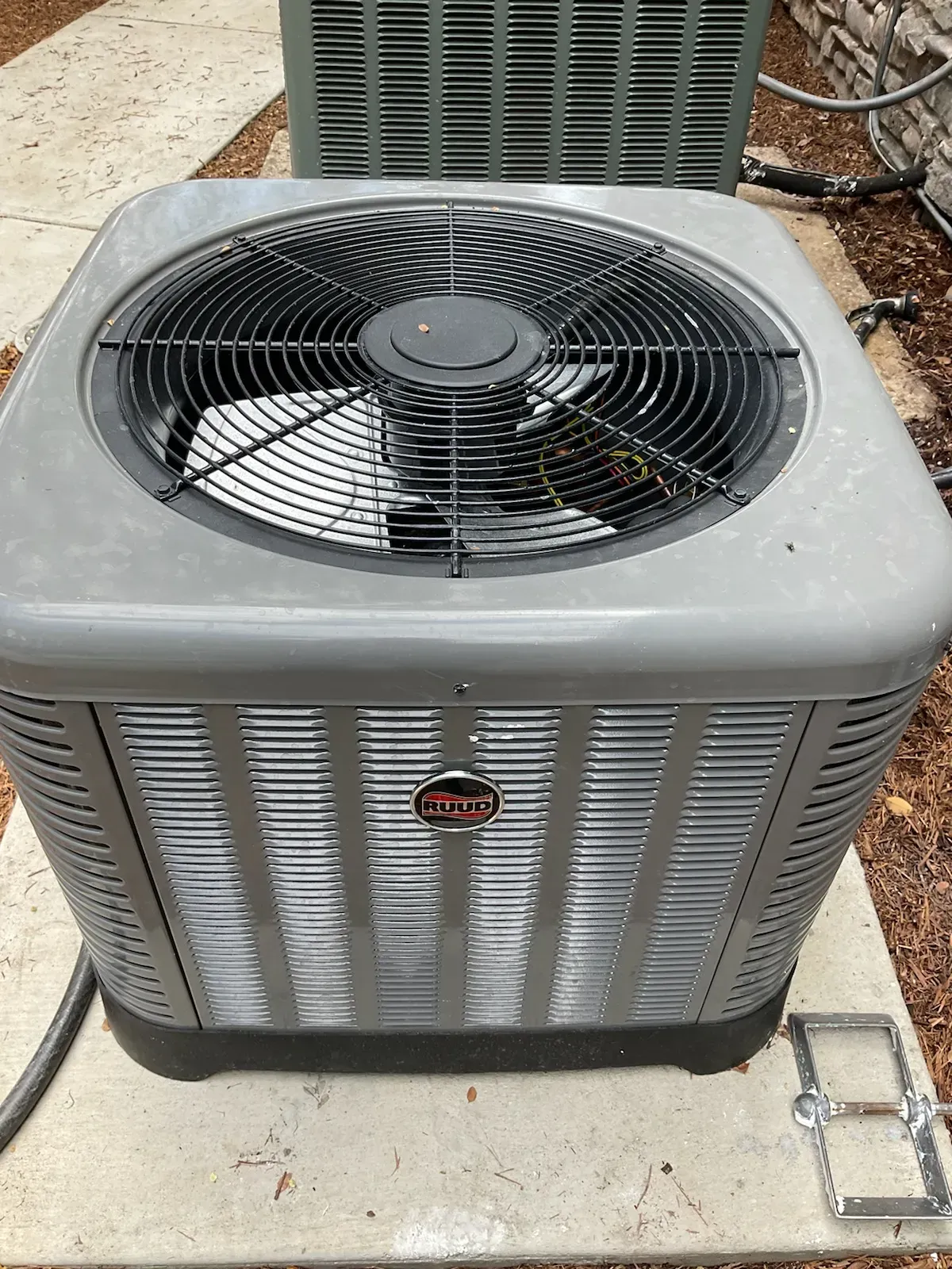 A gray air conditioner is sitting on a sidewalk next to a brick wall.