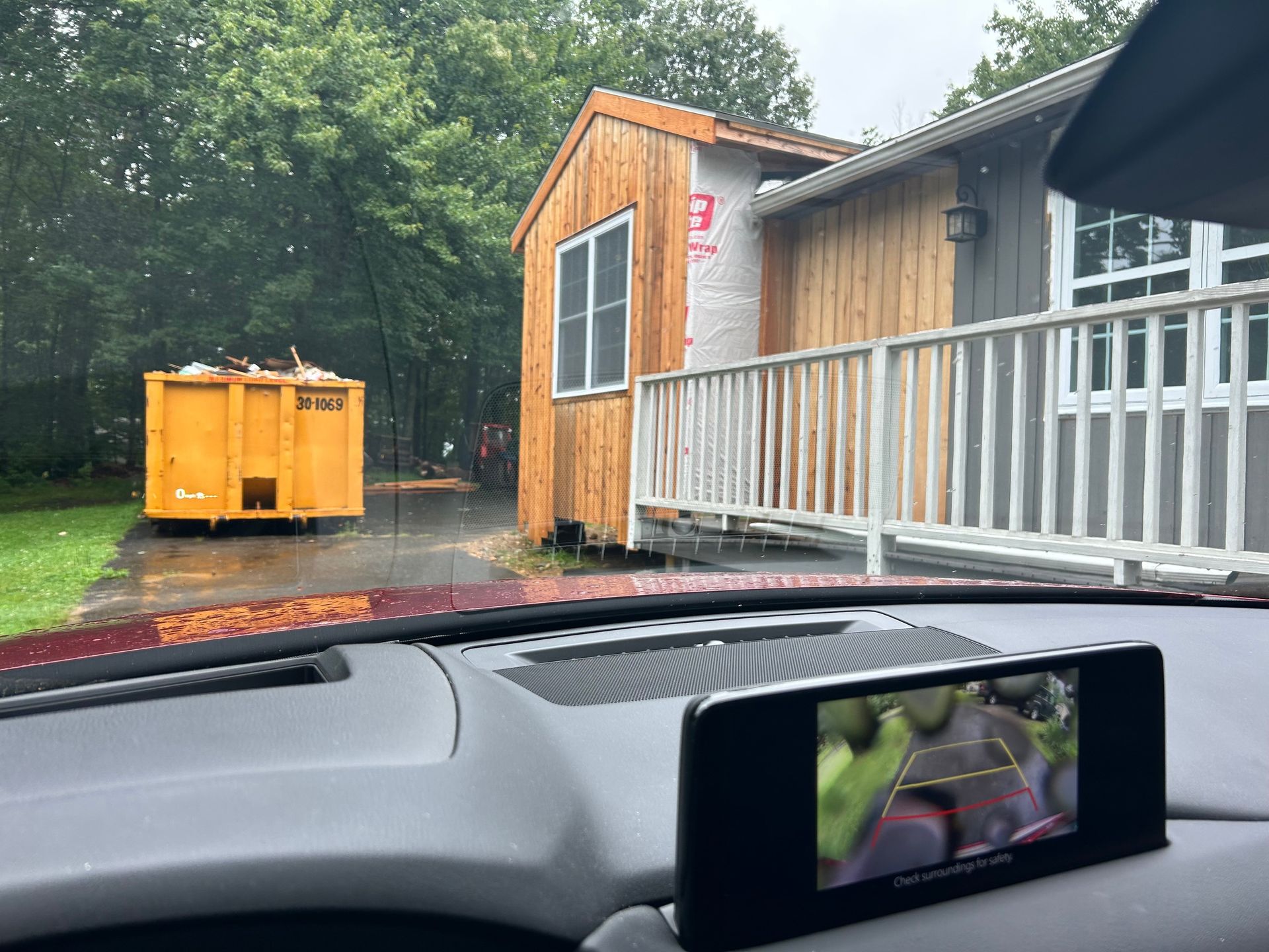 A car is parked in front of a house with a dumpster in the background.