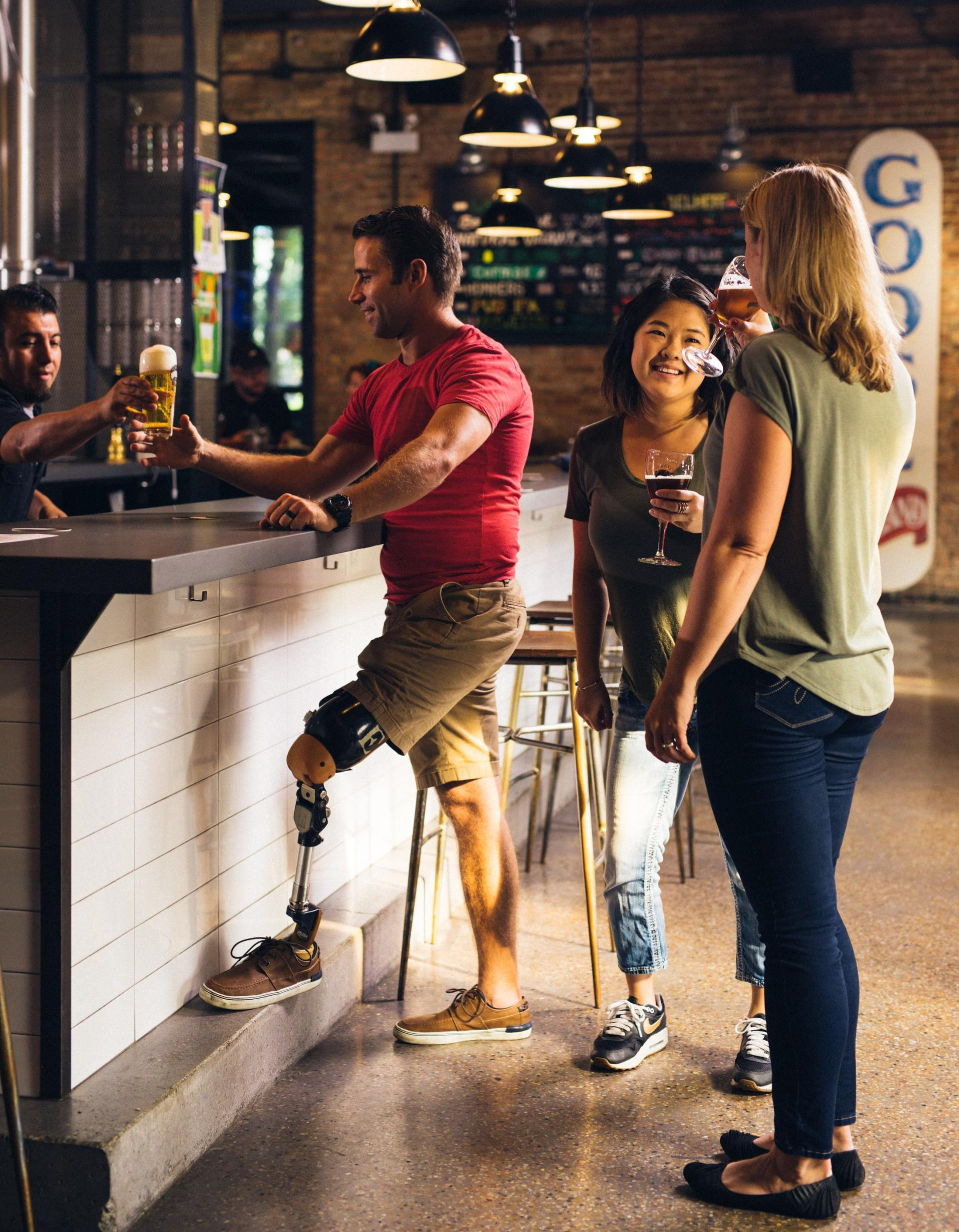 A man with a prosthetic leg is standing at a bar talking to two women.