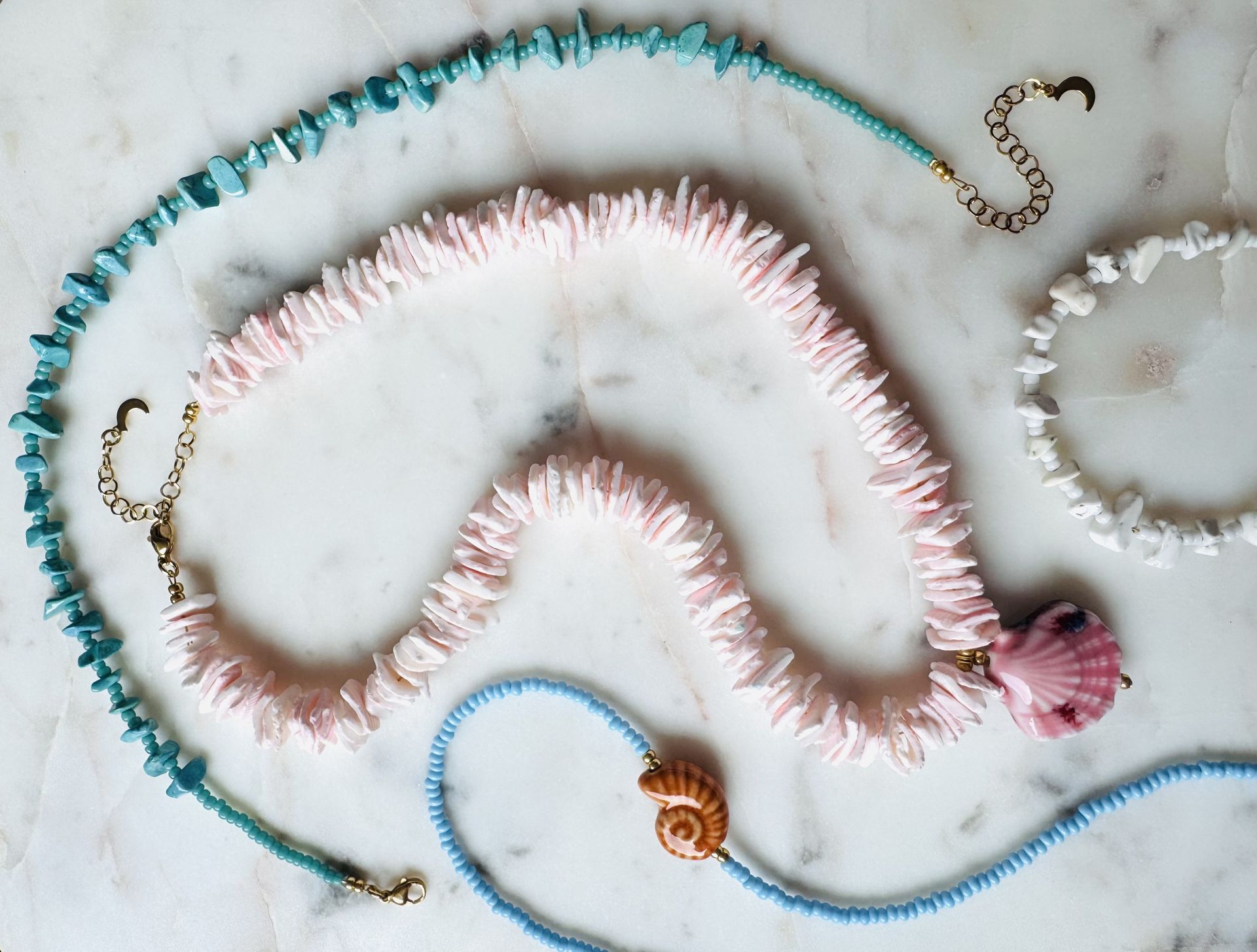 Vier kettingen van schelpen in roze, blauwe en witte tinten, gerangschikt op een marmeren ondergrond.