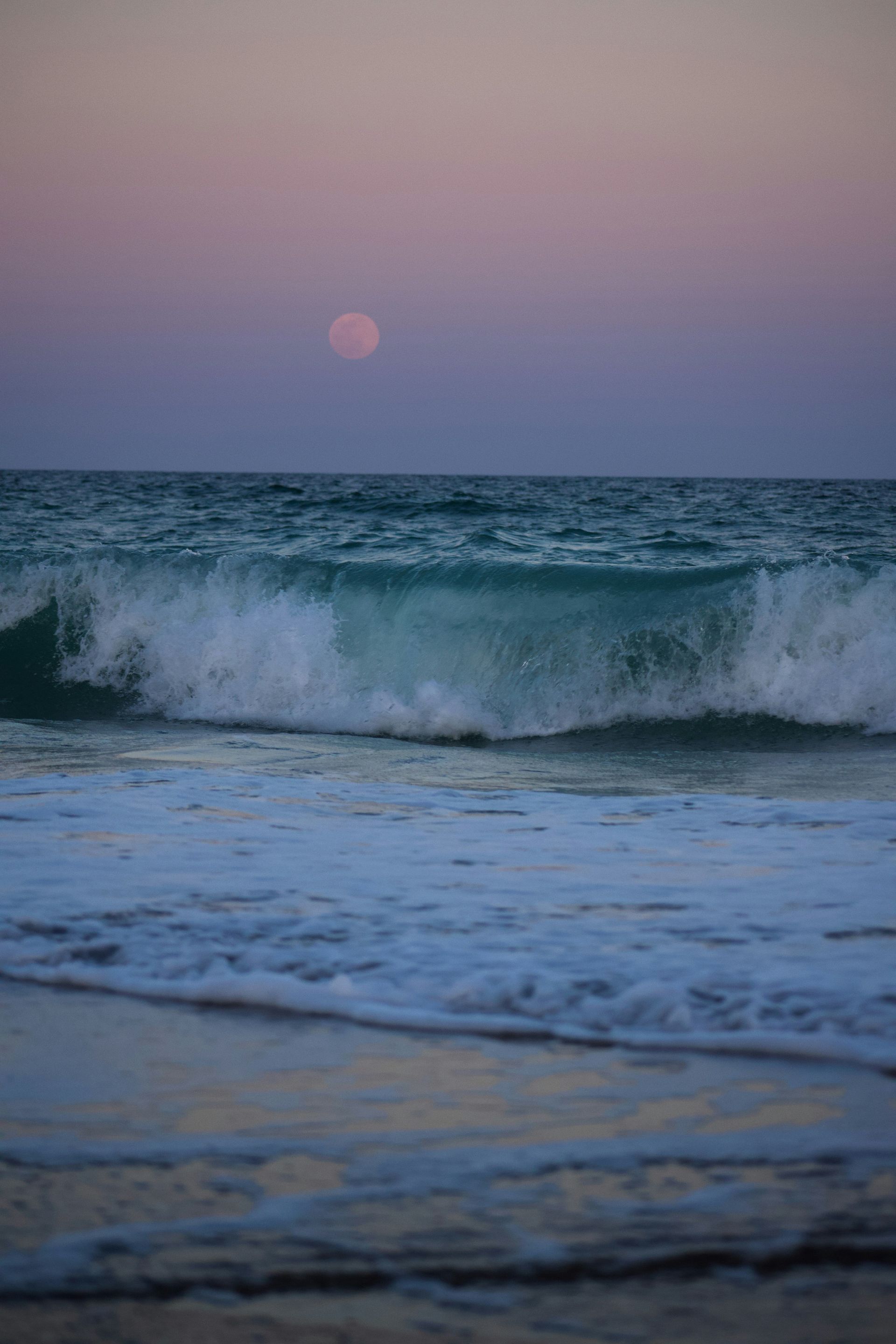 A full moon is rising over the ocean with waves crashing on the beach.