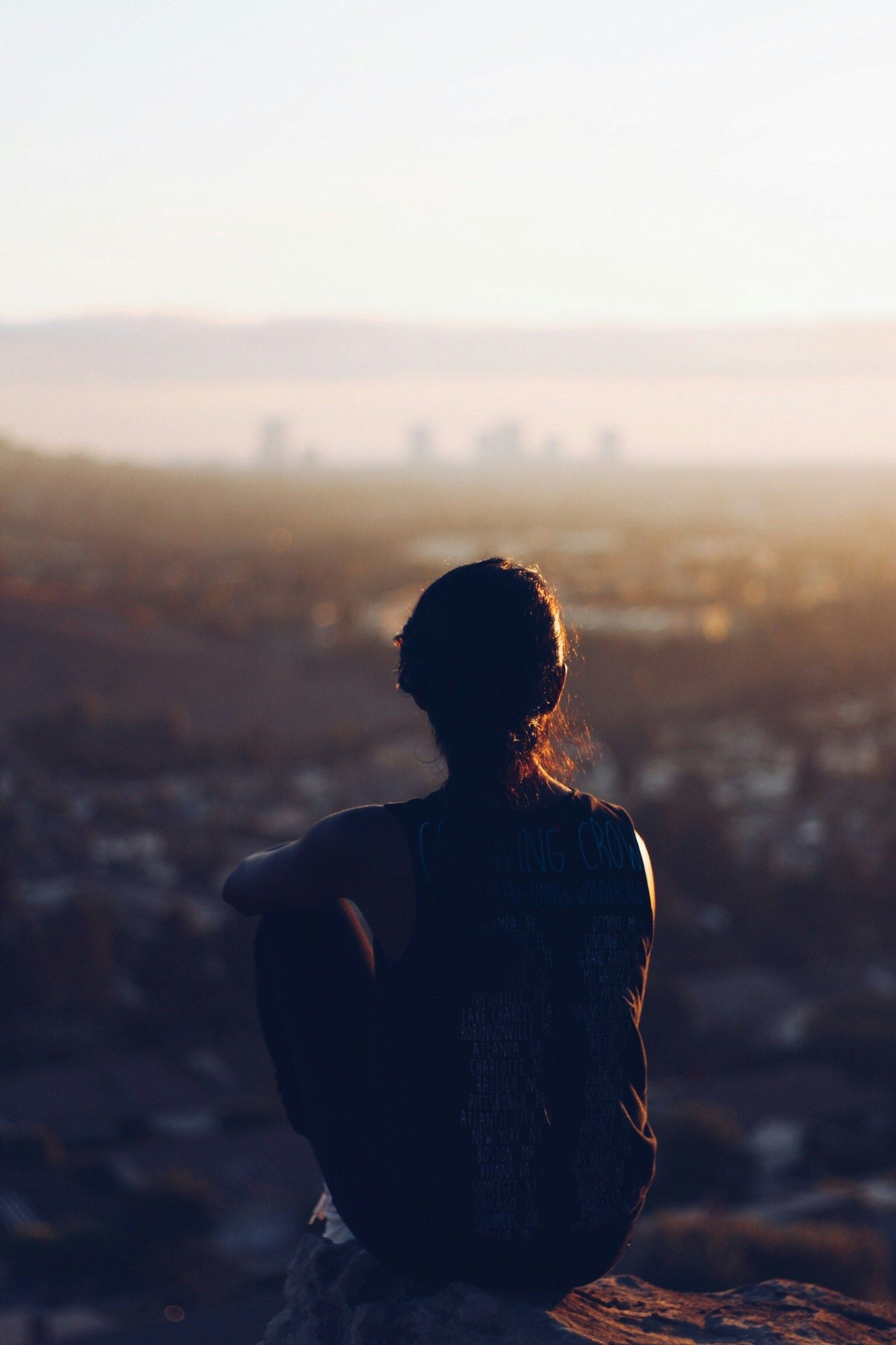 A woman is sitting on a rock overlooking a city at sunset.