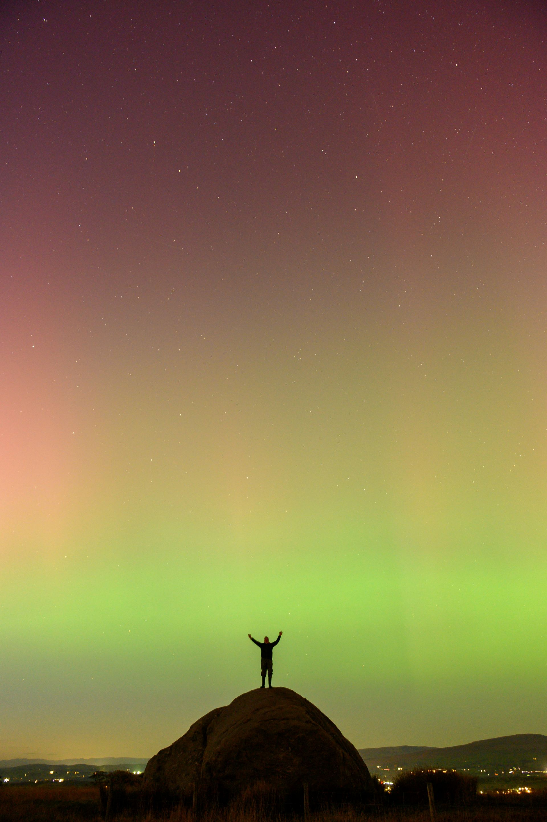 A person is standing on top of a hill in front of the aurora borealis.
