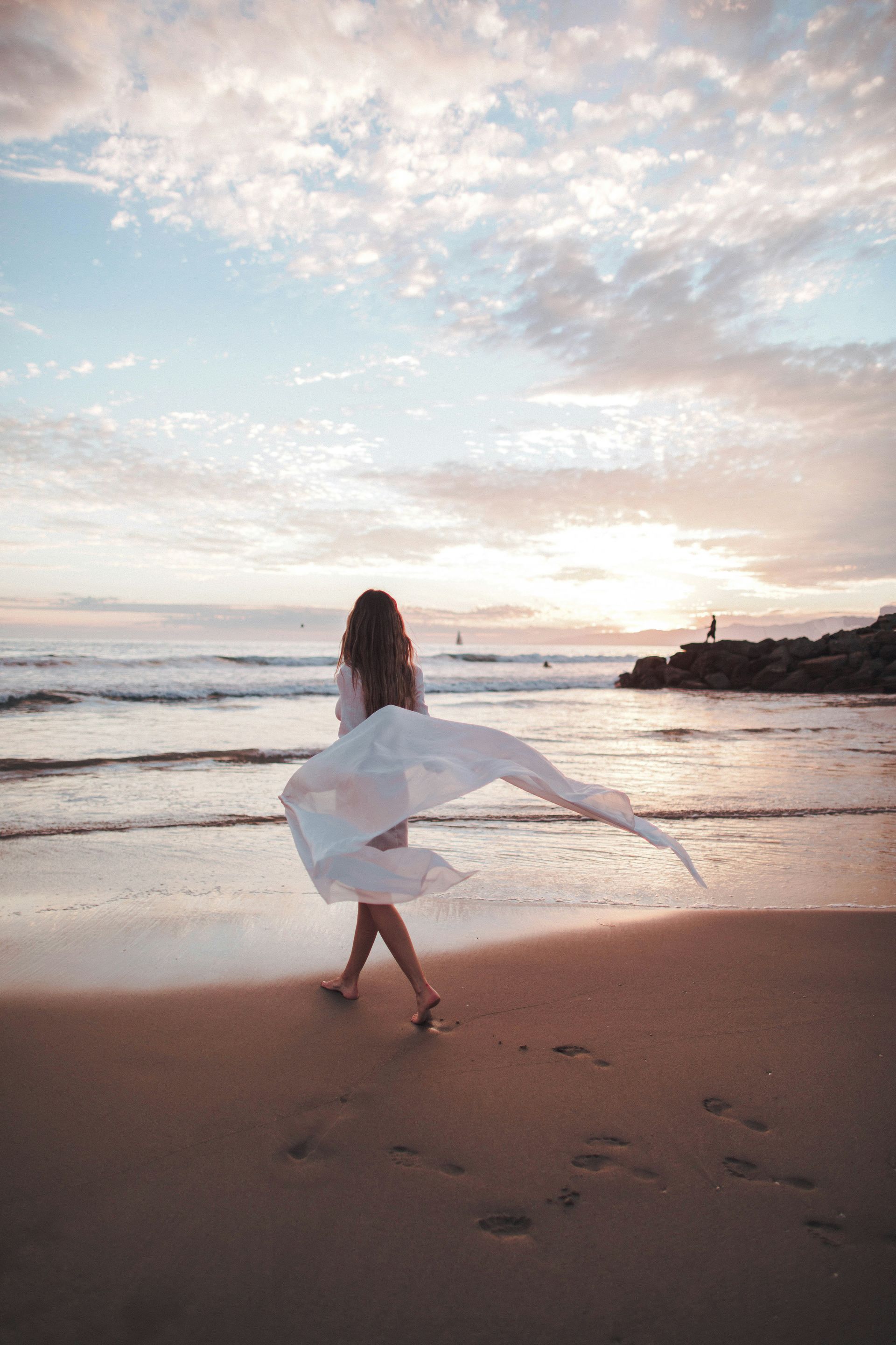 A woman in a white dress is walking on the beach at sunset.