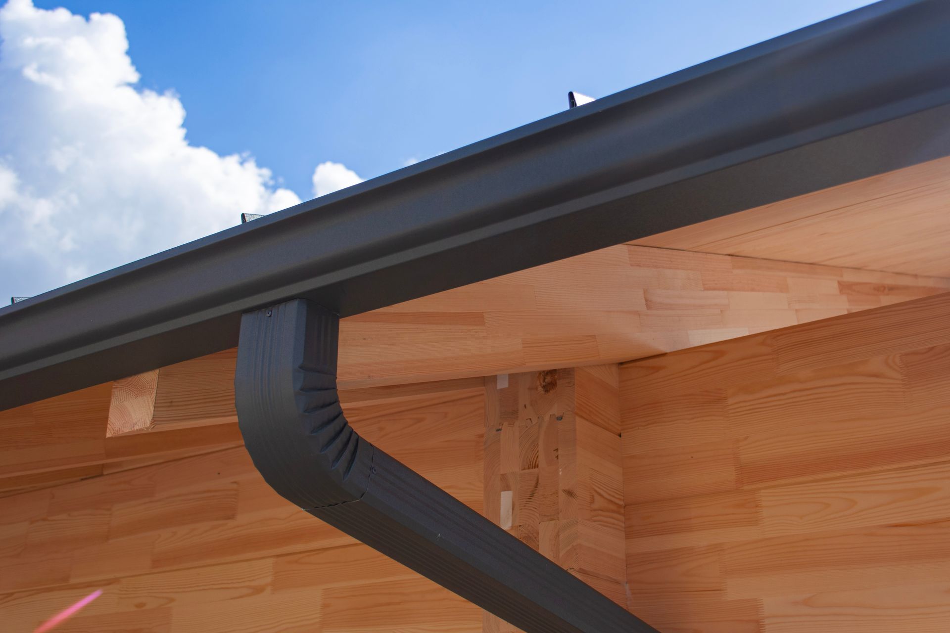 Dark gray rain gutter and downspout on a wood-paneled building with a blue sky background.