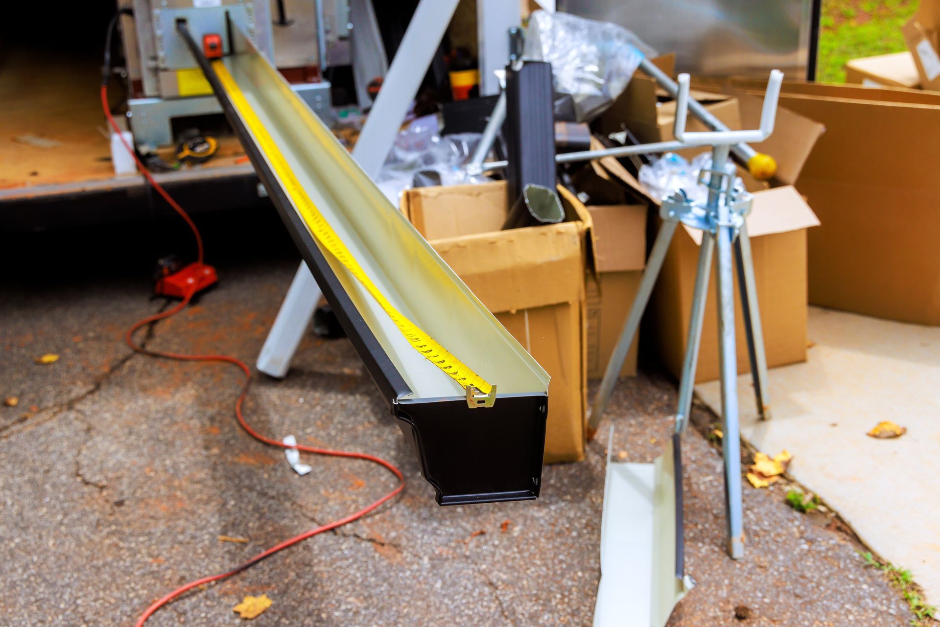 Black gutter with a yellow measuring tape inside, sitting on a work surface. Brown boxes and tools are in the background.