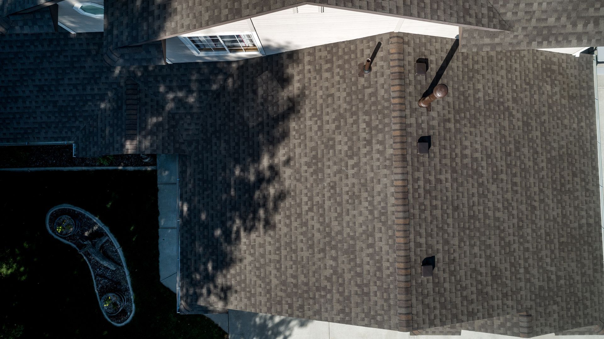 Overhead view of a house roof with a chimney and vents, cast in shadow from a nearby tree.