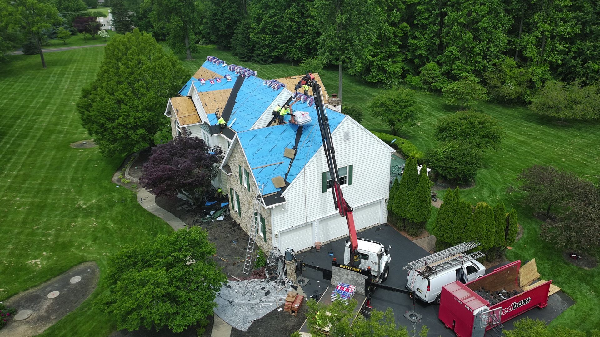 Roof being replaced on a two-story house, with workers and a crane, surrounded by green trees and grass.