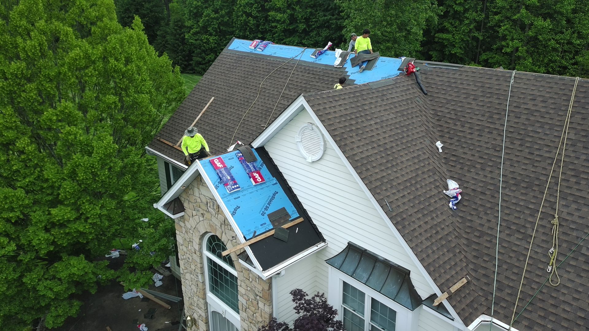 Roofers replacing shingles on a house roof. Workers in yellow vests. Blue underlayment visible. Green trees in the background.