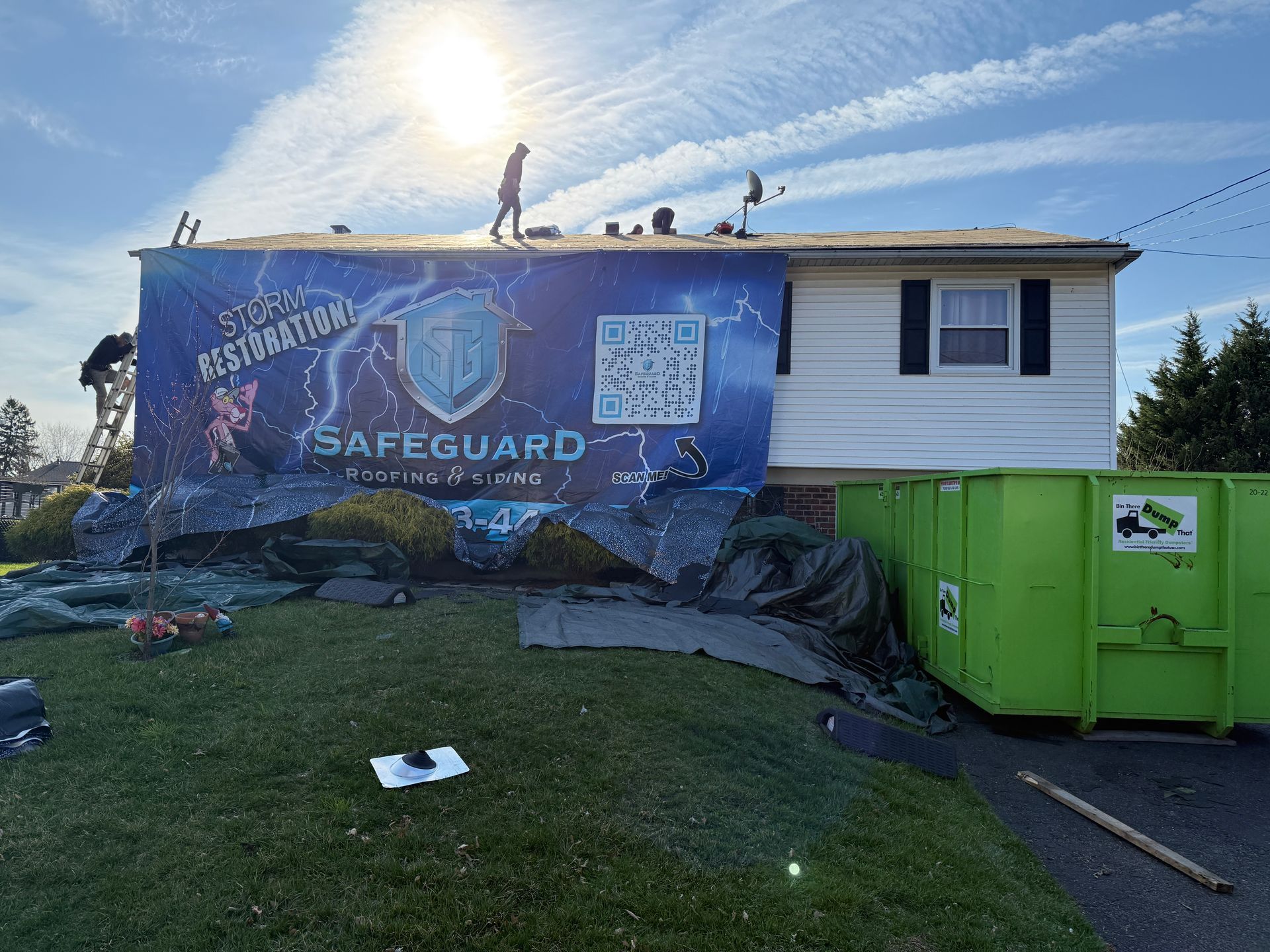 Roofing workers on a house with a large banner. Green dumpster on right. Bright sun.