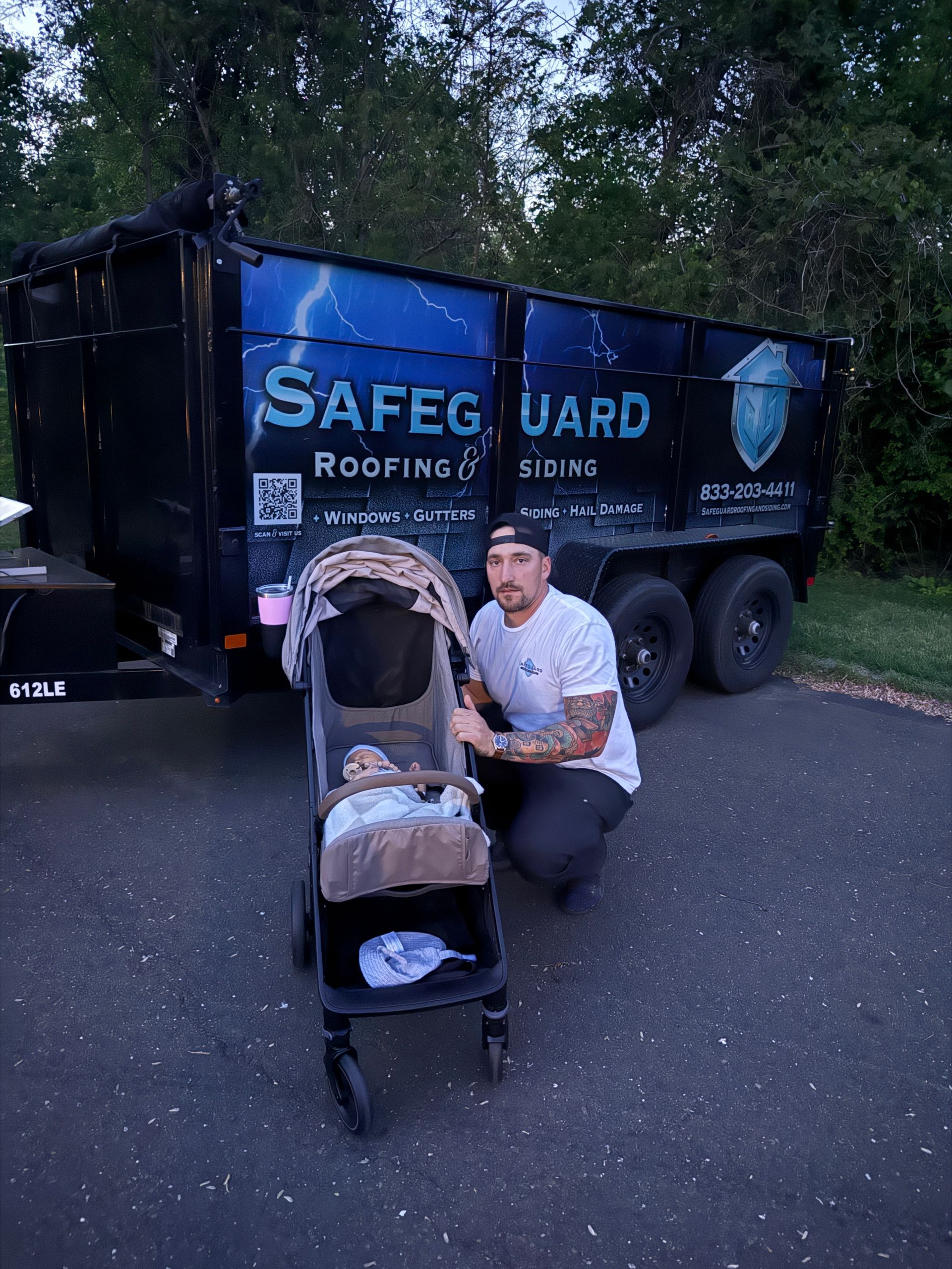 Man kneels beside a stroller, in front of a dumpster with the logo for a roofing company on it.