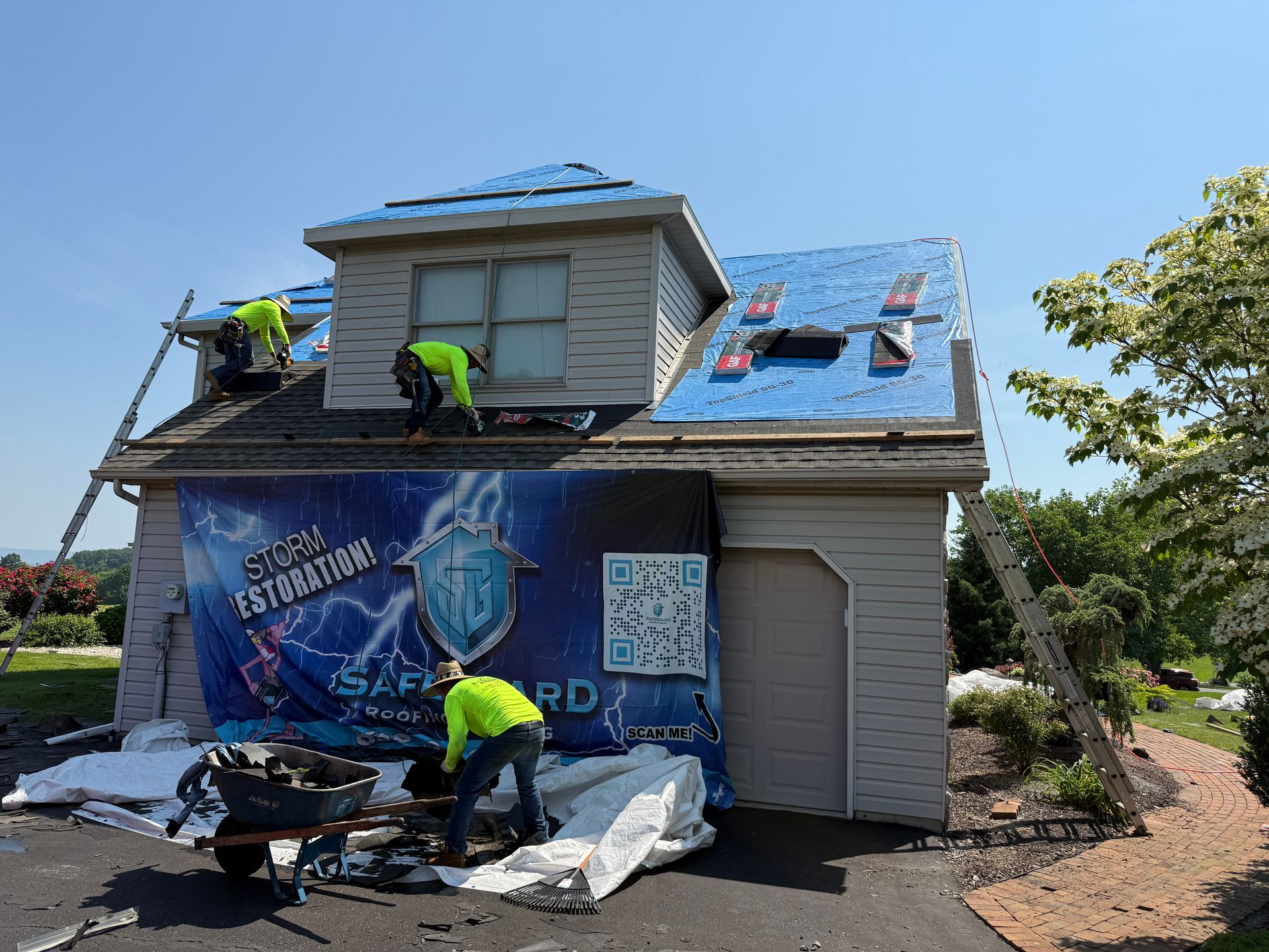 Roofers replacing shingles on a house; men working, blue tarp, sunny day.