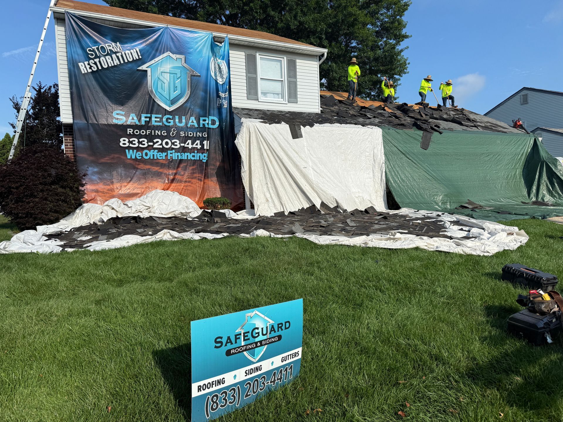 Roof damage covered with tarps. Workers in yellow vests on the roof. Safeguard company sign on lawn.