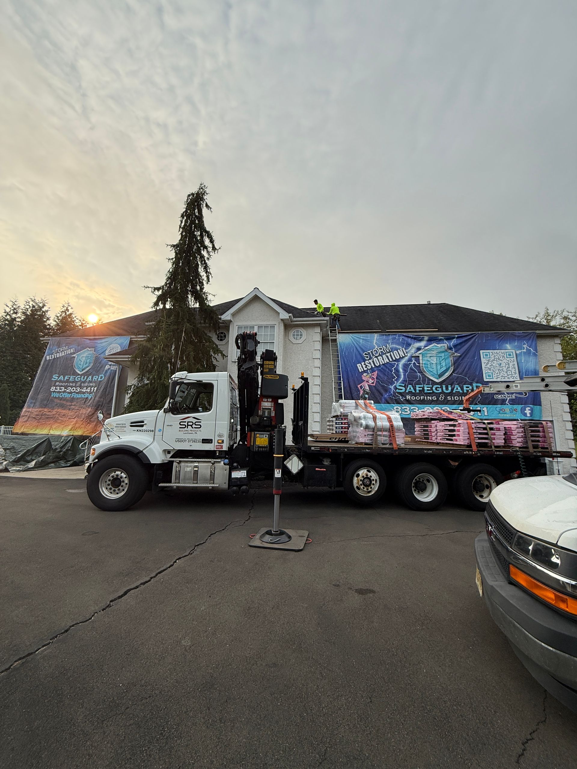 Truck with crane parked in front of a house, loaded with roofing materials. Large banners with logo visible.