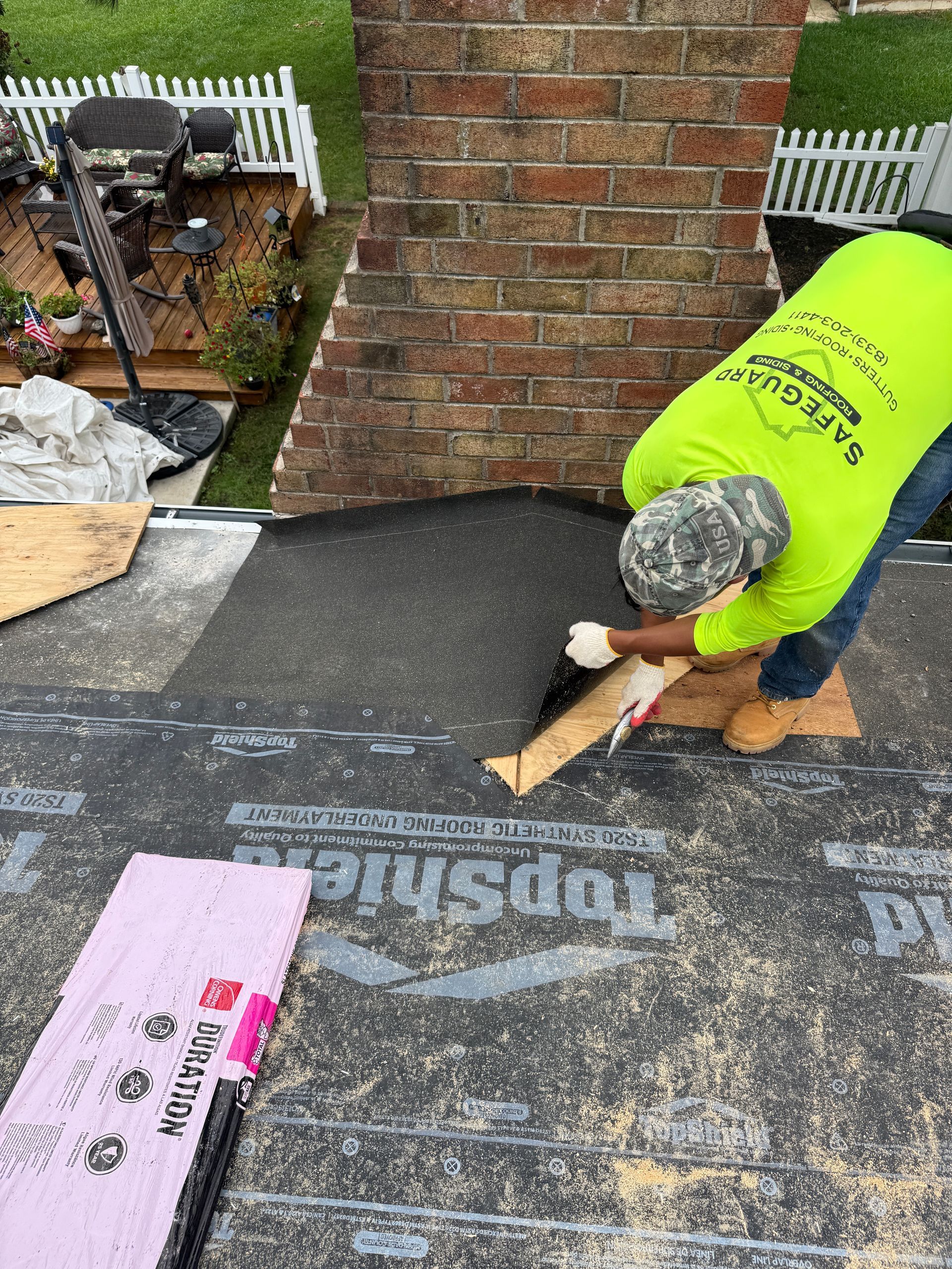 Roofer in safety vest applying sealant near a brick chimney on a flat roof, surrounded by materials.