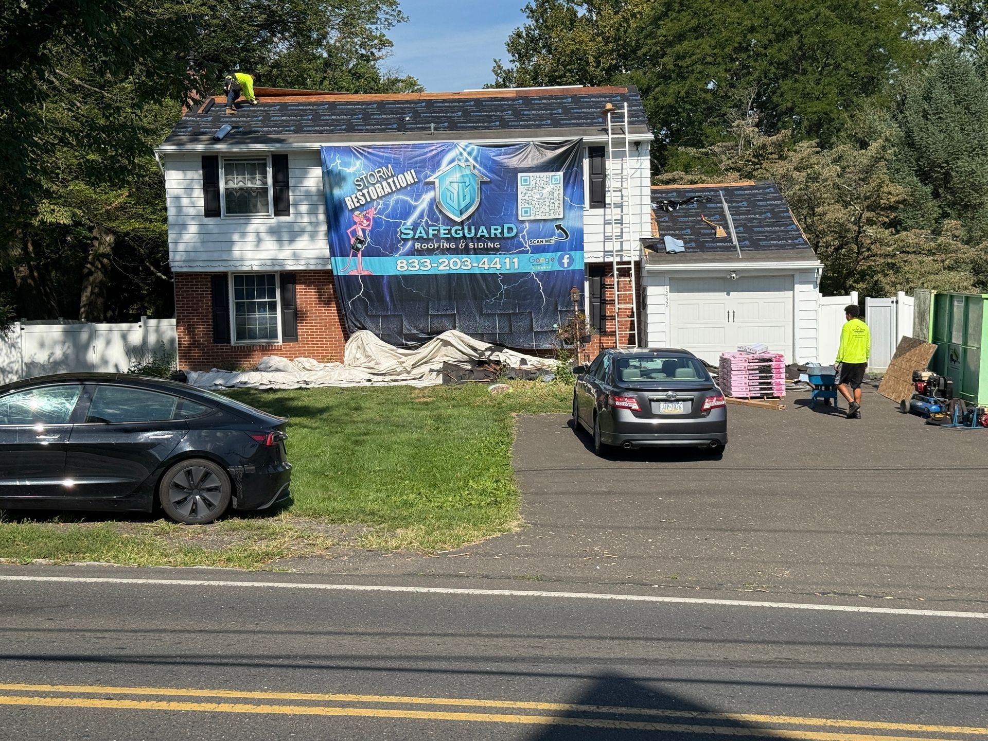 House roof being repaired; black tarp and sign, cars in driveway, worker on roof.