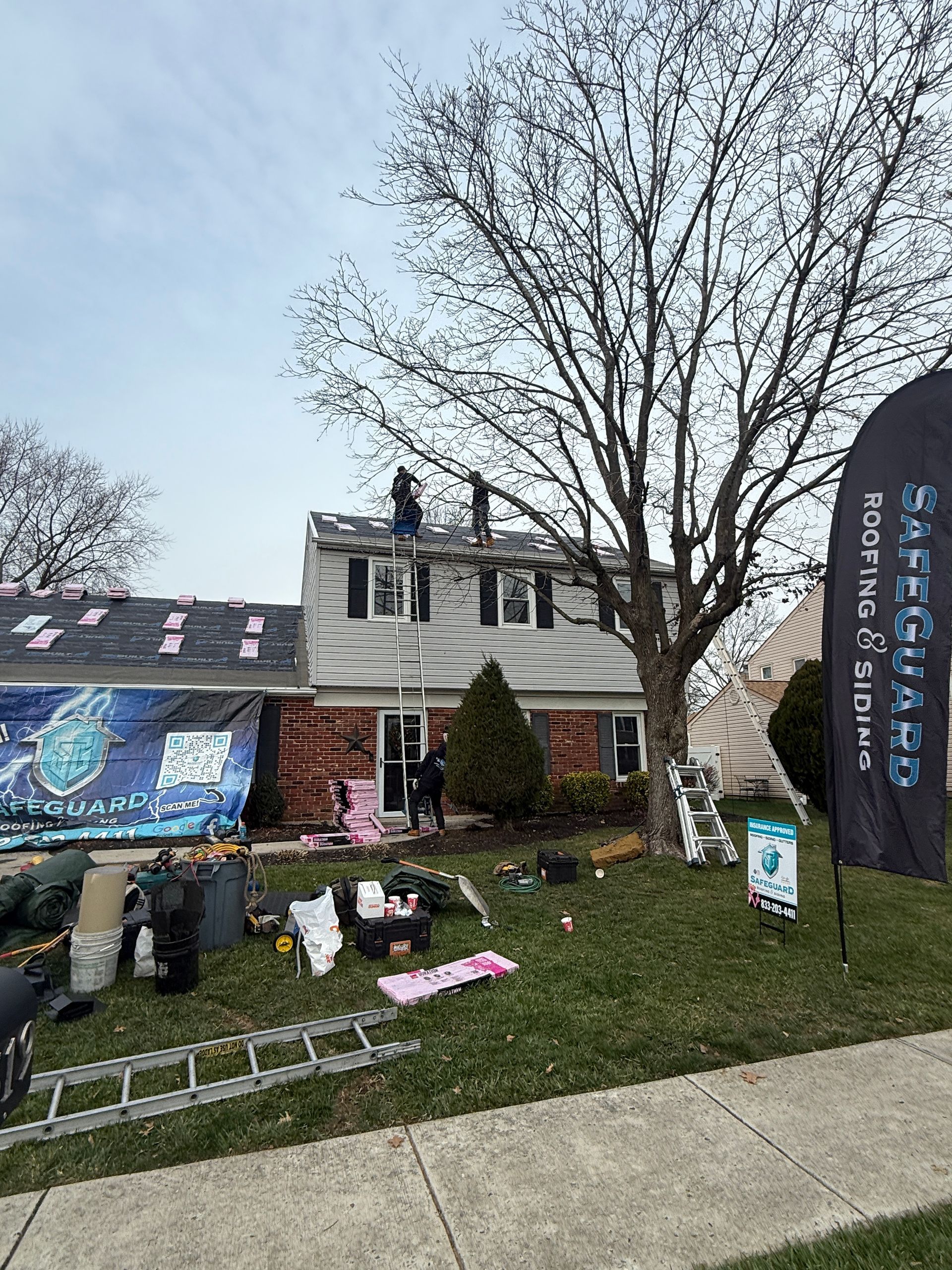 Roofers working on a two-story house with a partially replaced roof. Equipment and sign in yard. Overcast sky.